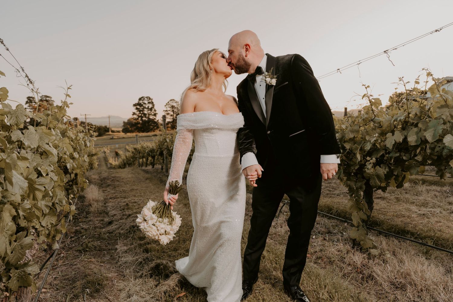 A newlywed couple walking through a vineyard at sunset wearing formal wedding attire.