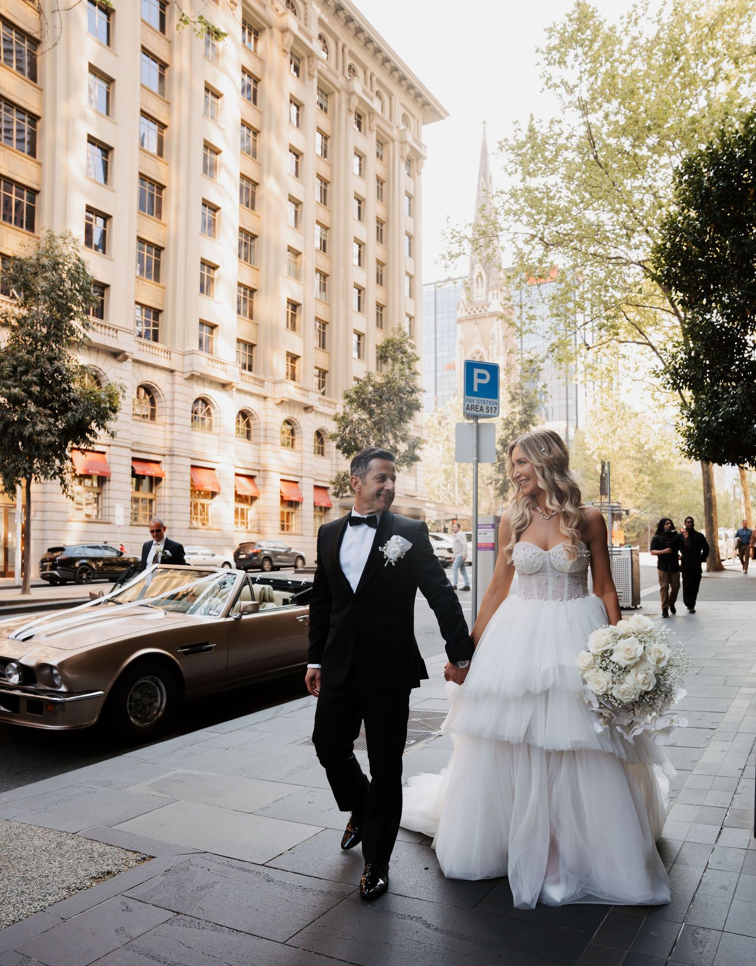 A couple celebrates their wedding day walking together on a city street with classic architecture and a vintage car.