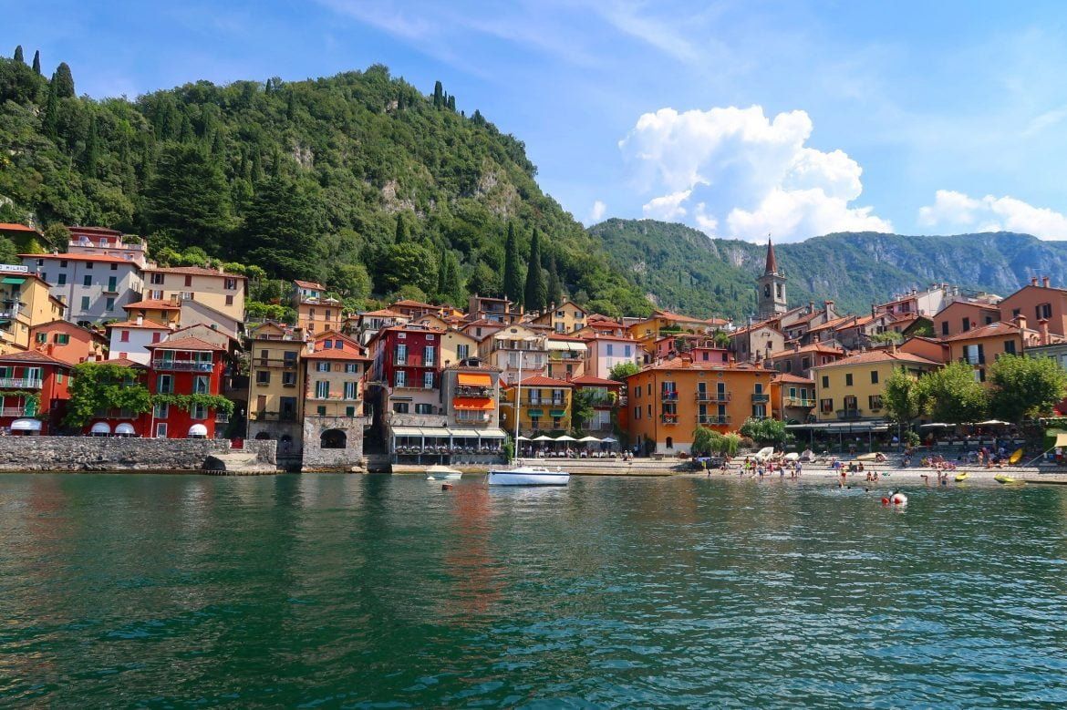Colorful Mediterranean houses line the waterfront of Lake Como in Varenna, Italy with mountains and boats in view.