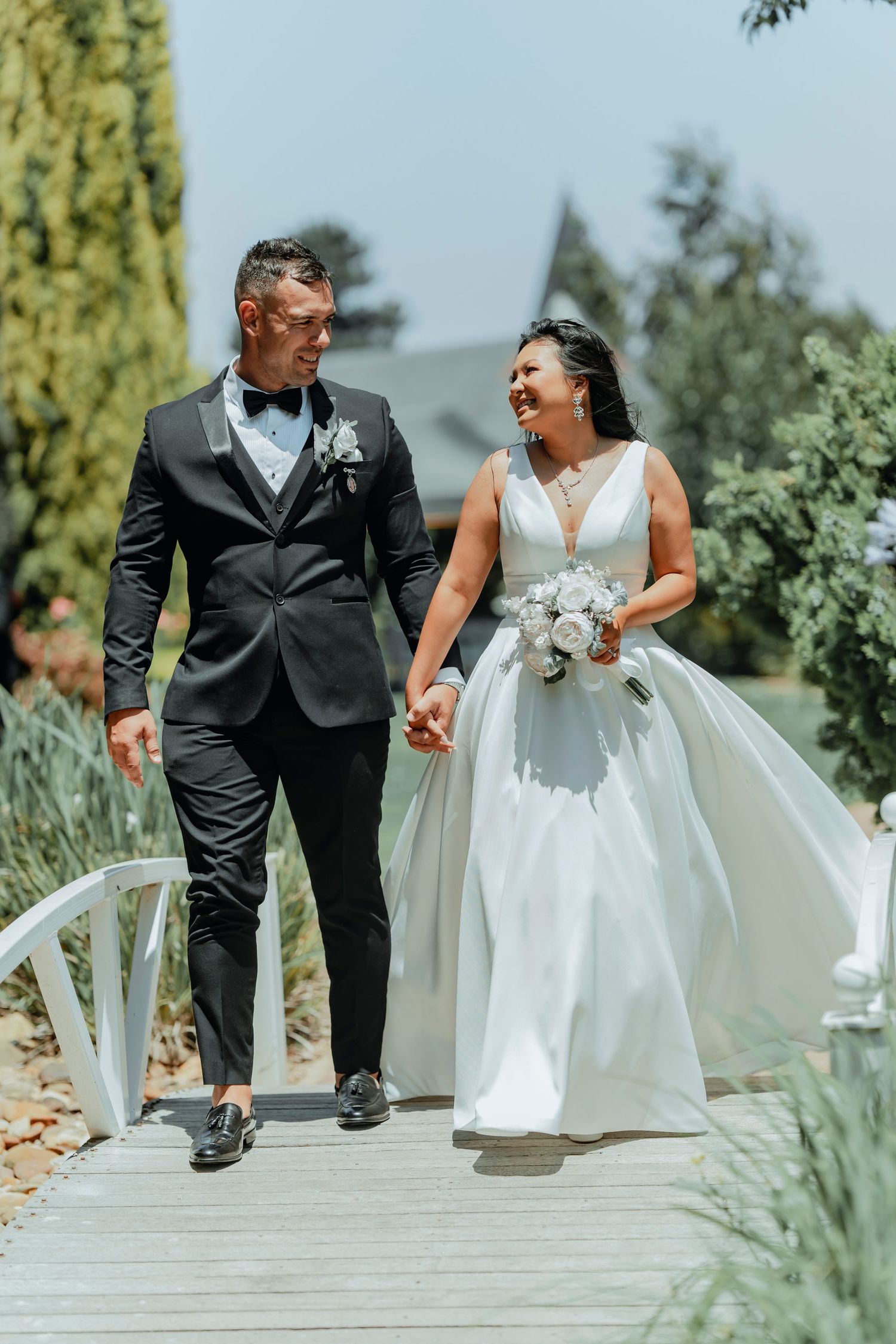 A bride in a white gown and groom in a black tuxedo walking together and laughing on a garden pathway.