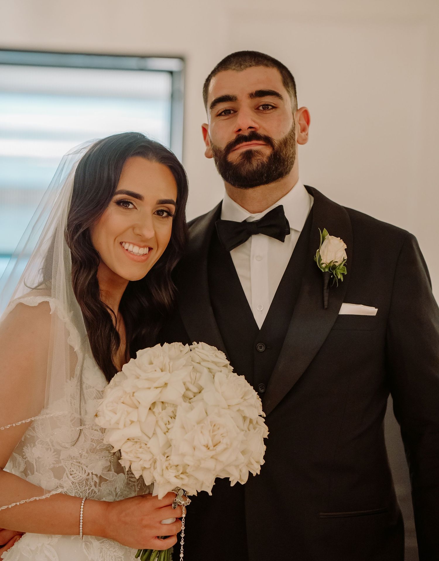A couple on their wedding day poses together in formal attire with a white hydrangea bouquet.
