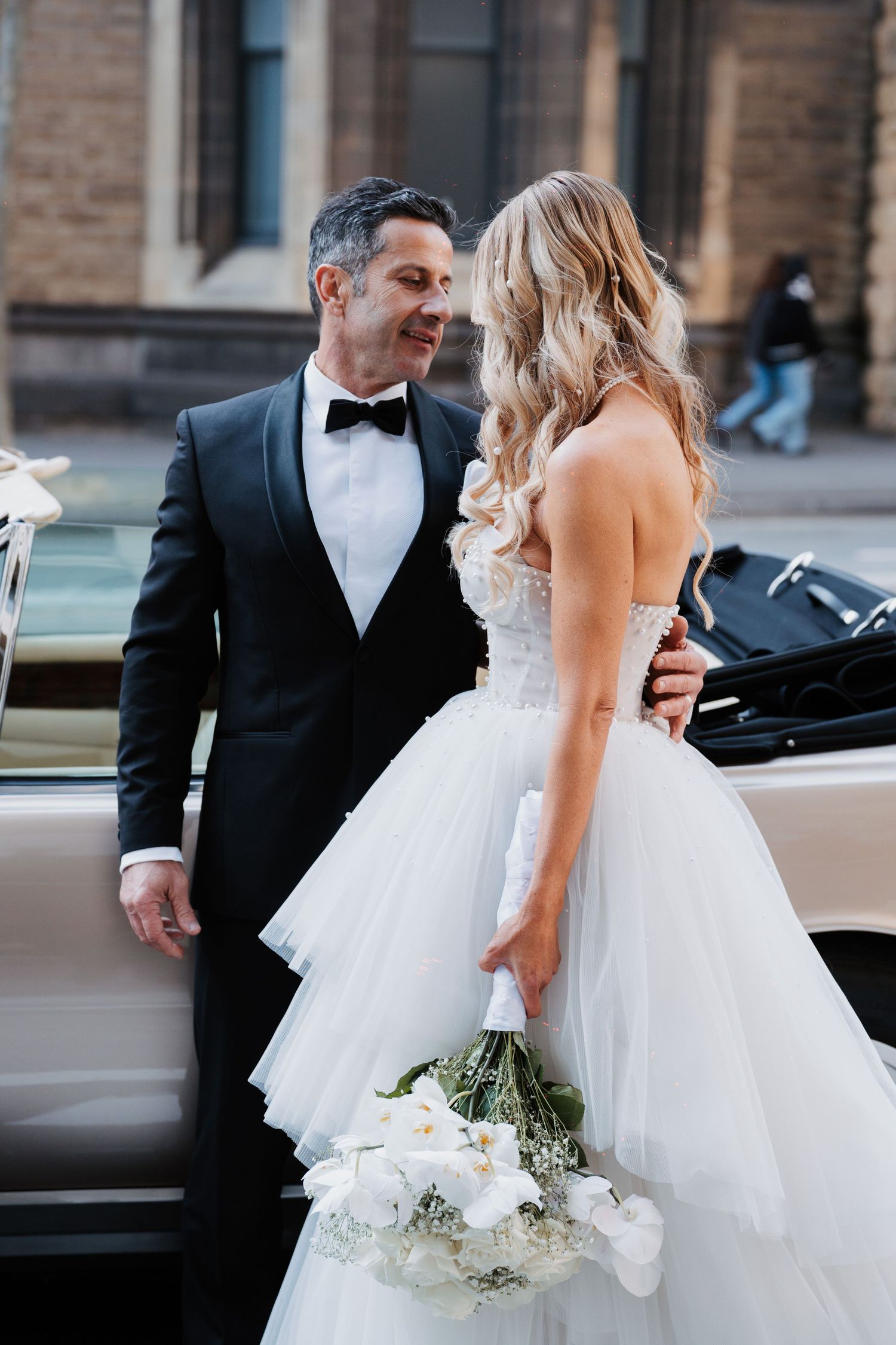 A romantic wedding moment between a couple by a vintage car, with the bride holding a white floral bouquet.