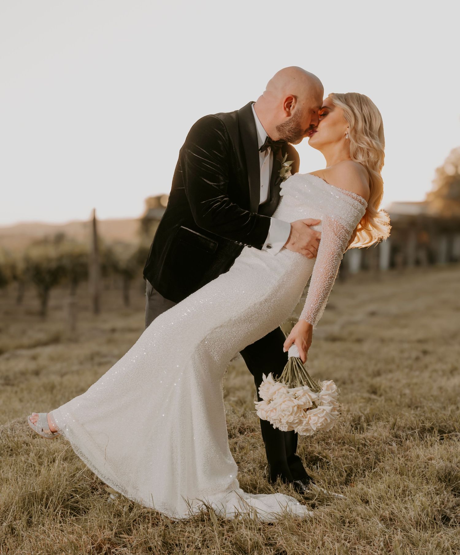 A romantic wedding couple shares a kiss in a vineyard at sunset while the bride dips back gracefully in her fitted white gown.