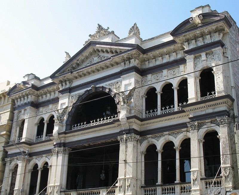 Historic colonial architecture with ornate white facade featuring classical columns and arched balconies in deteriorating condition.
