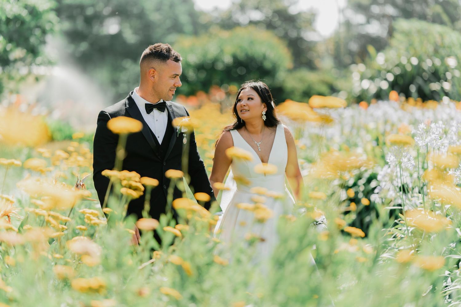 A couple stands together in a dreamy garden filled with yellow wildflowers and soft sunlight on their wedding day.