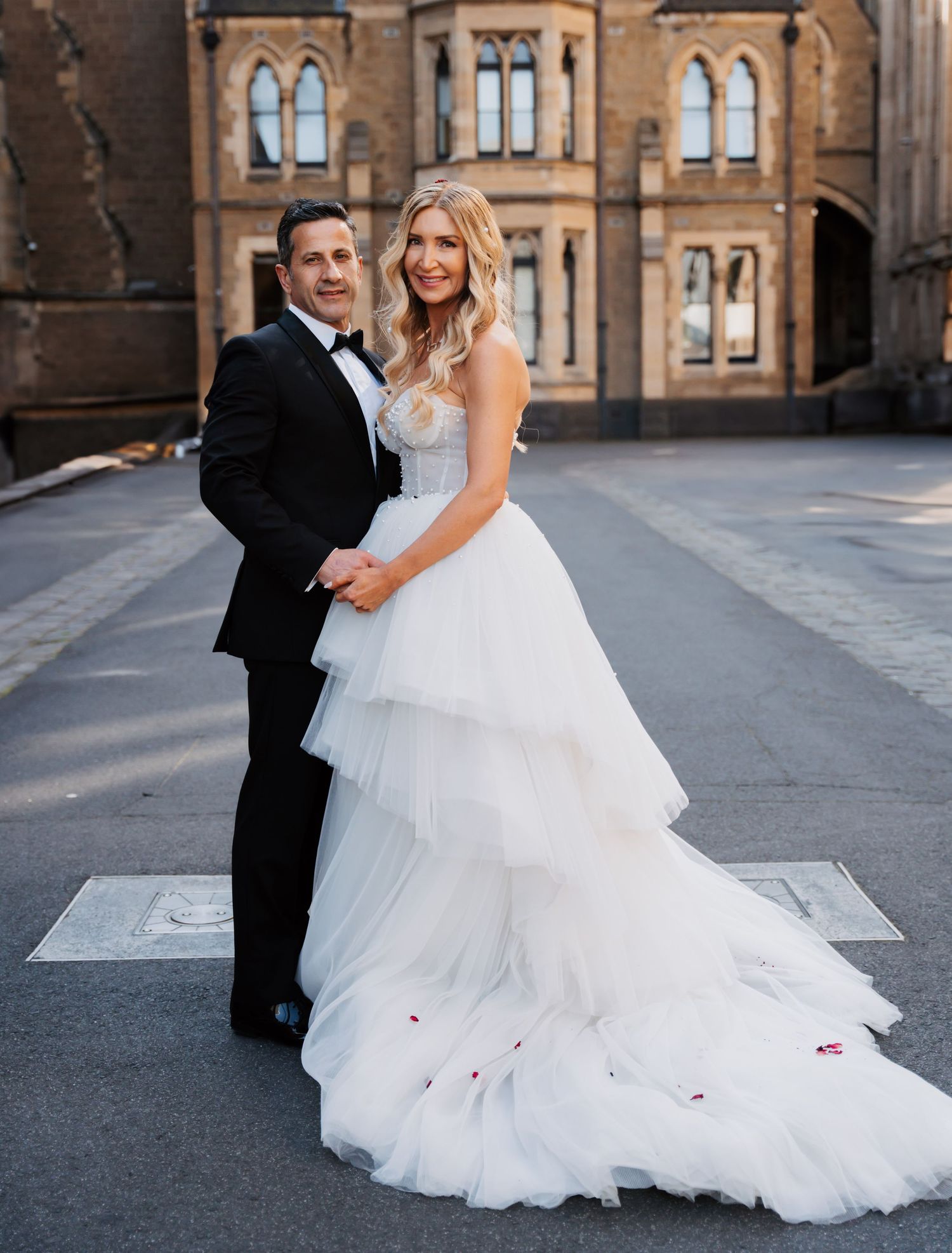 A couple poses in formal wedding attire outside a historic stone building with Gothic architecture and arched windows.