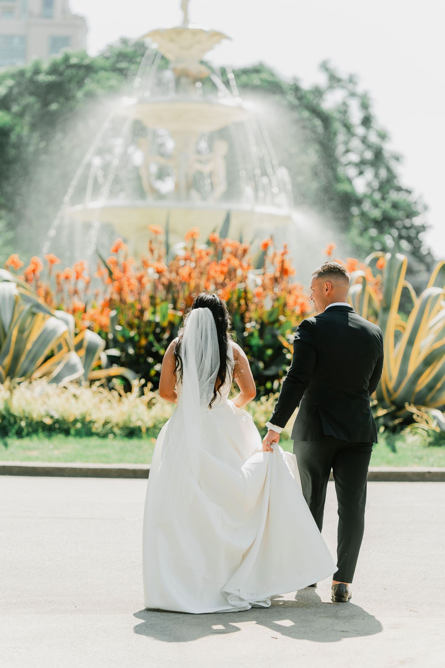 A newlywed couple walks toward an ornate fountain surrounded by vibrant orange flowers and lush garden landscaping.