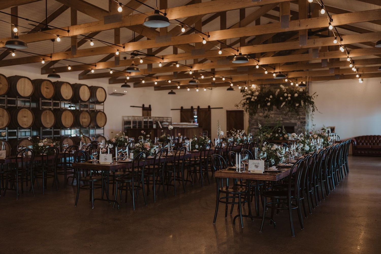 A rustic wine barrel room set up for an elegant reception with long tables, string lights, and wooden ceiling beams.