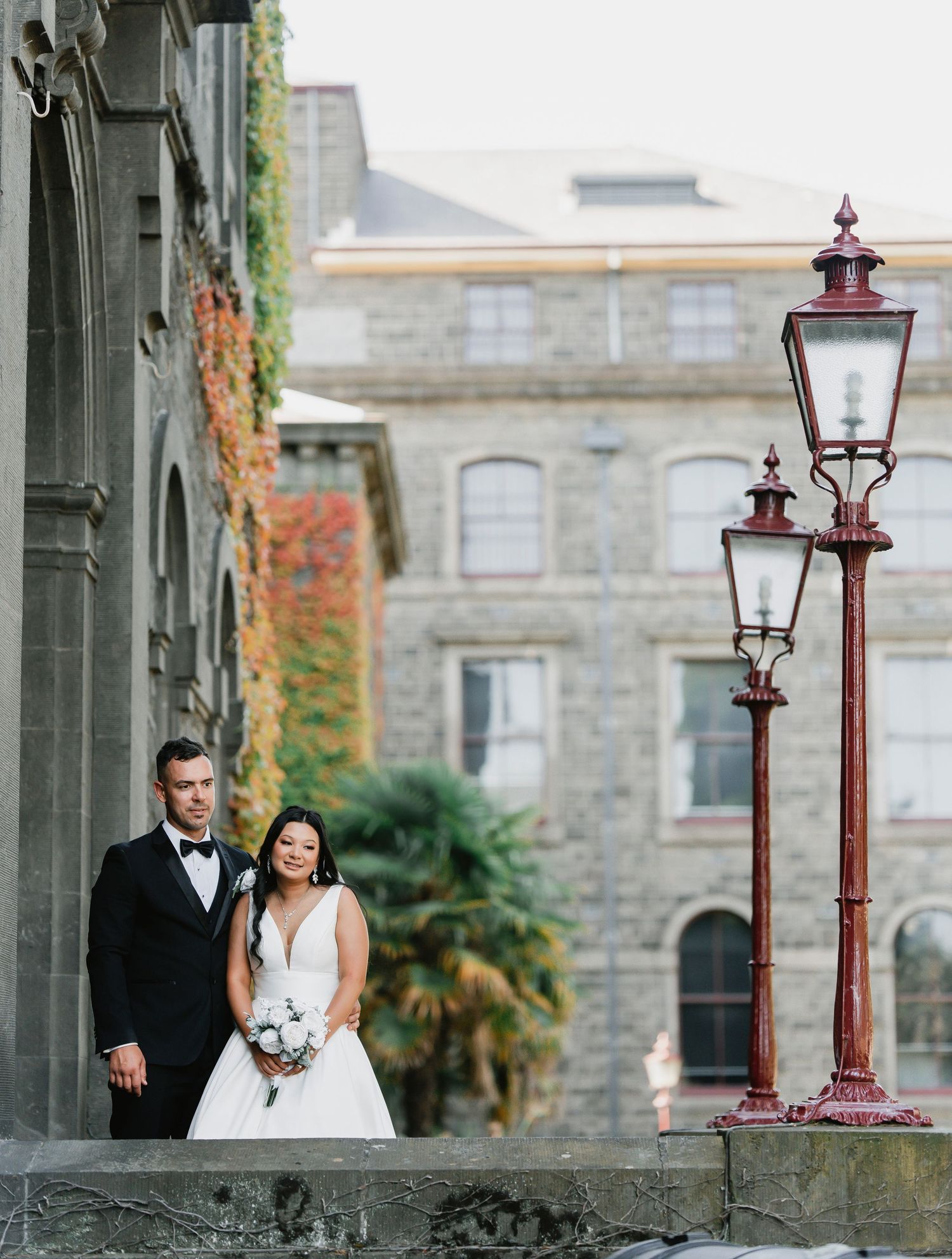 Elegant couple pose beside vintage lampposts on stone steps with autumn ivy and historic building facade.