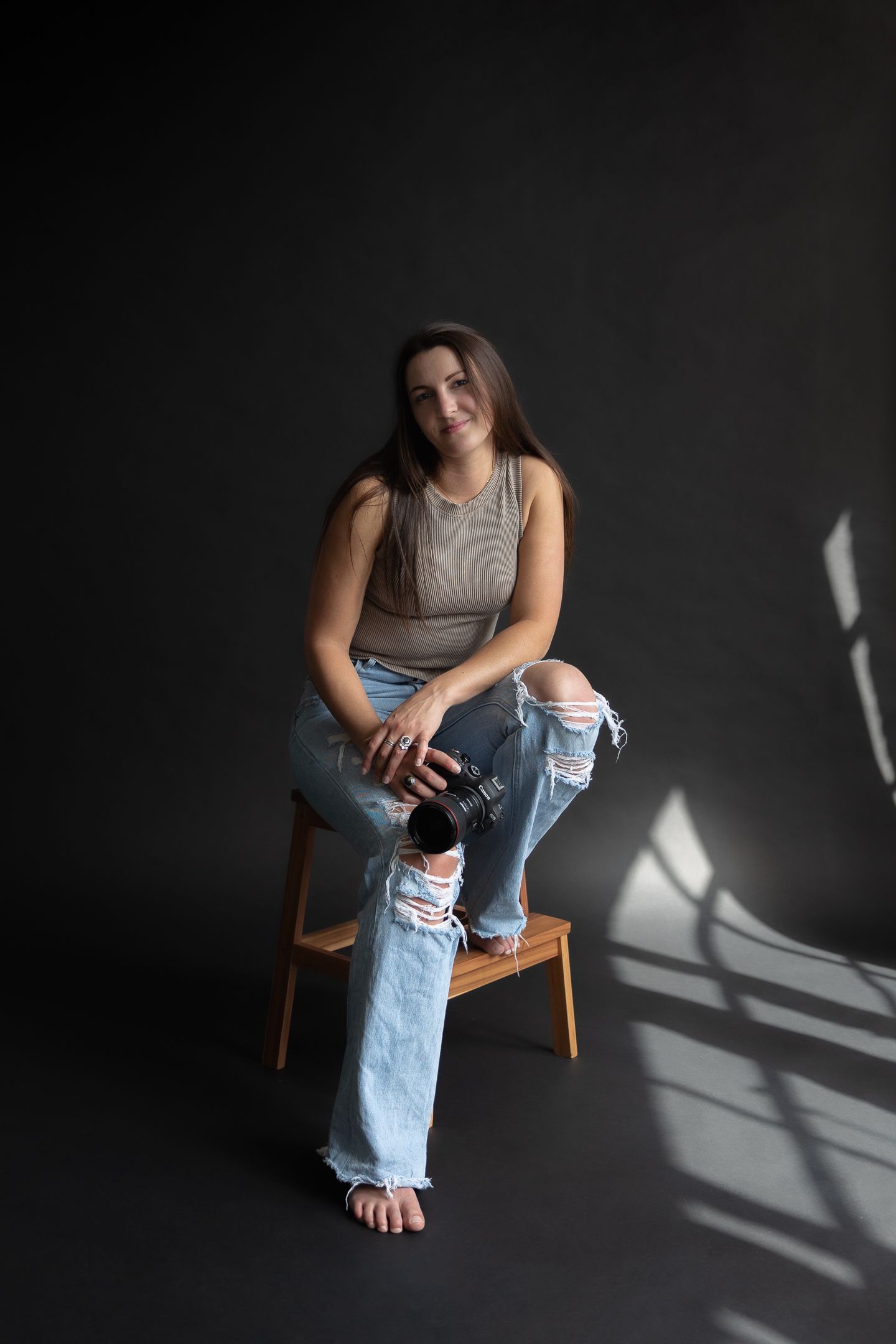 lady smirking at camera sitting on stool with camera in hand wearing brown tank and ripped jeans against black backdrop
