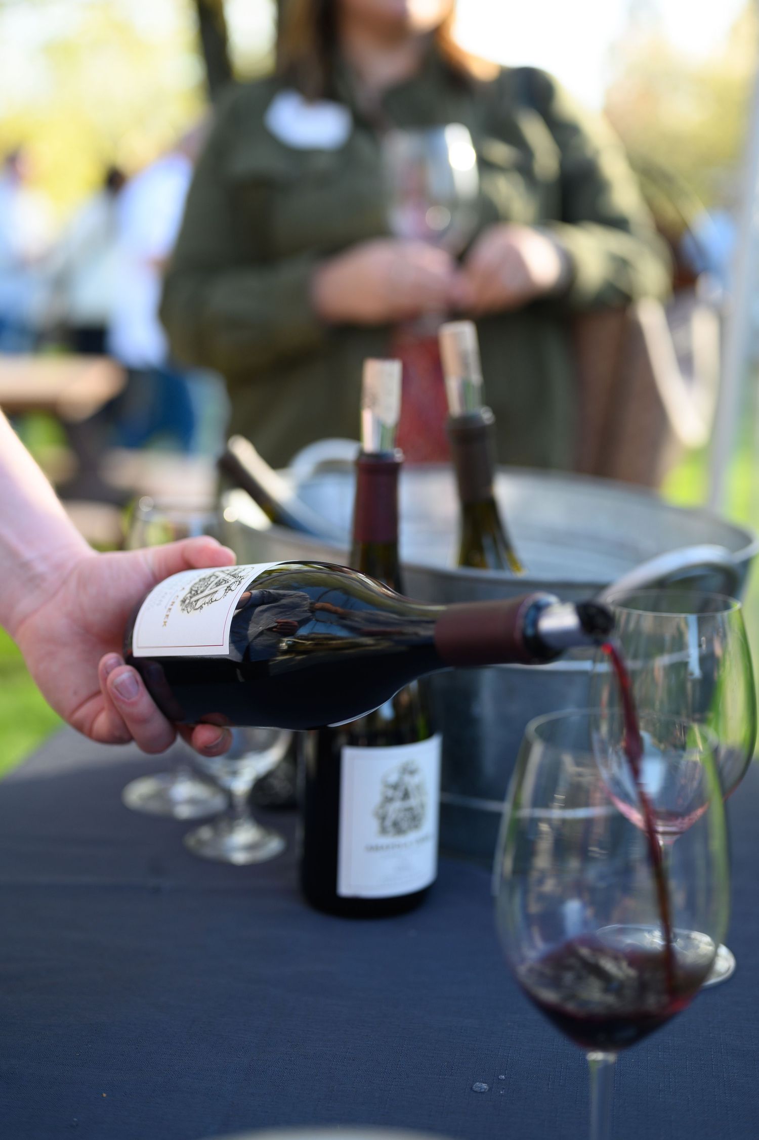 Someone pours red wine from a bottle into glasses during an outdoor wine tasting event.