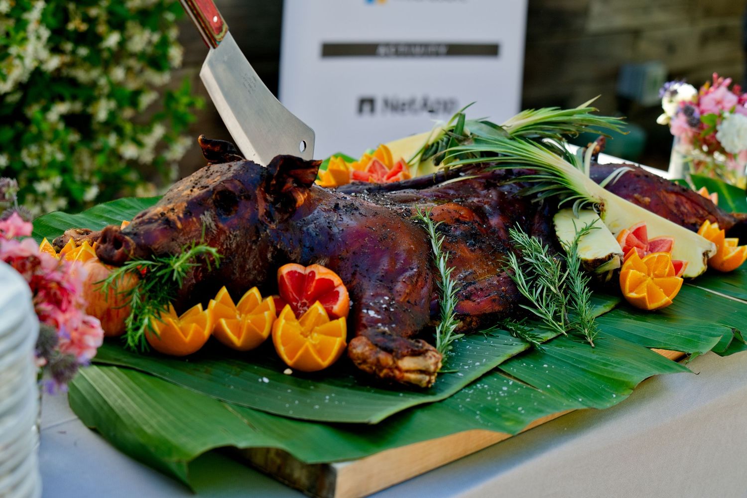 A roasted whole pig garnished with fresh herbs and orange flowers rests on green banana leaves at a festive luau.