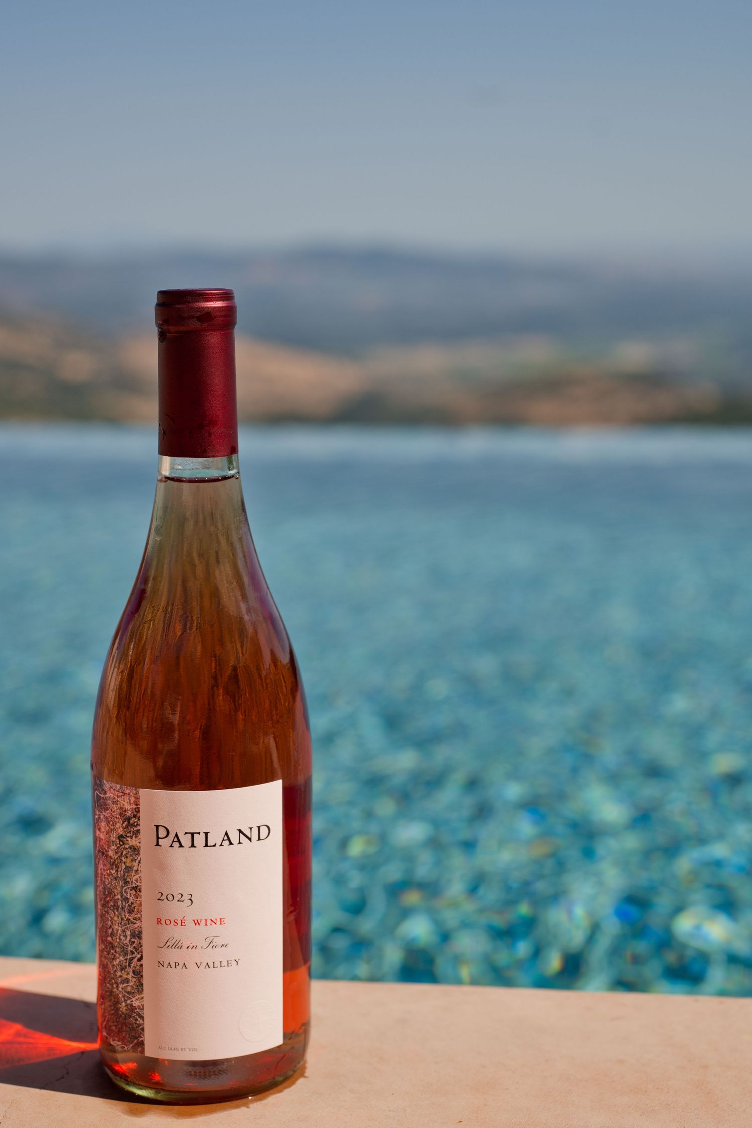 Bottle of Patland rose wine sits on pool ledge with scenic mountain vista, napa valley, and turquoise water in background.