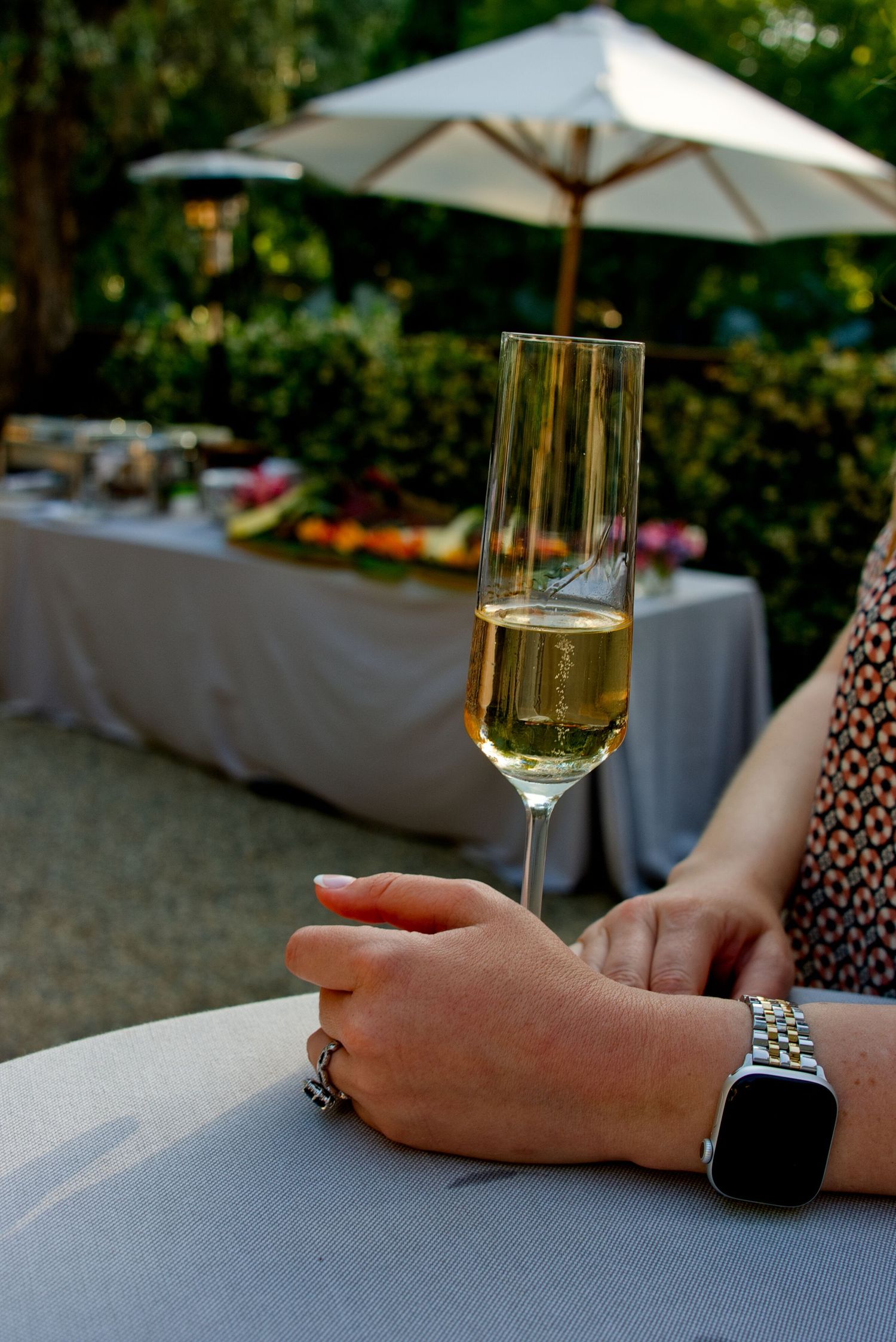A hand wearing a smartwatch holds a glass of sparkling wine at an outdoor event with white umbrellas and tables.