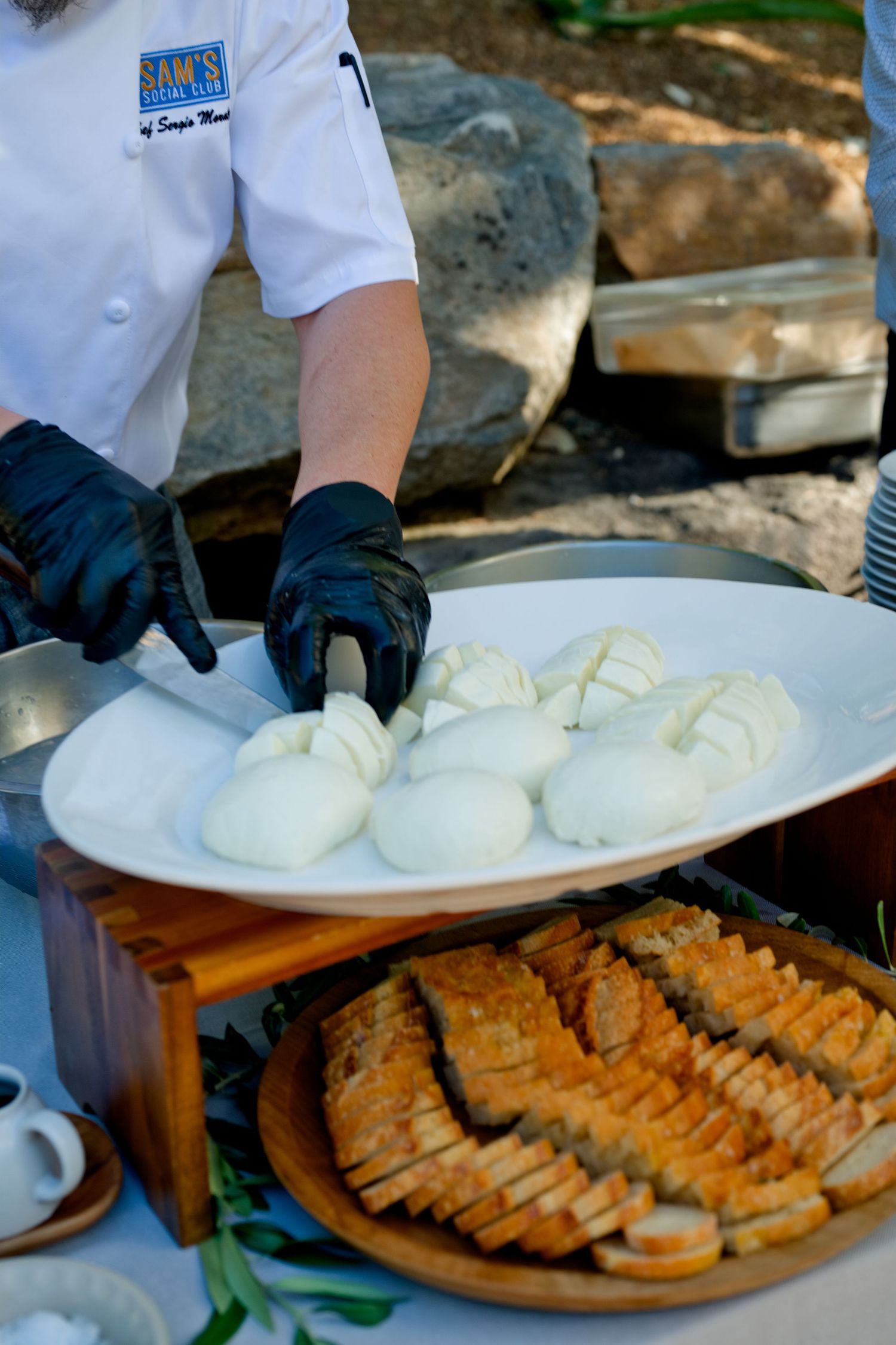 Chef wearing white uniform and black gloves plates fresh mozzarella alongside sliced bread at outdoor catering event.