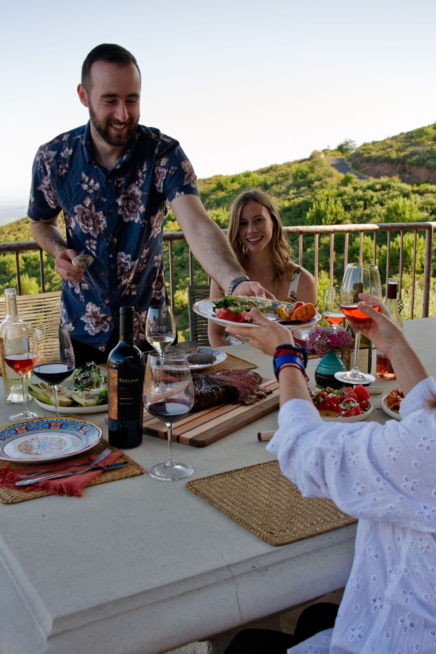Friends sharing food and wine at an outdoor dinner party on a balcony with scenic mountain views at sunset.