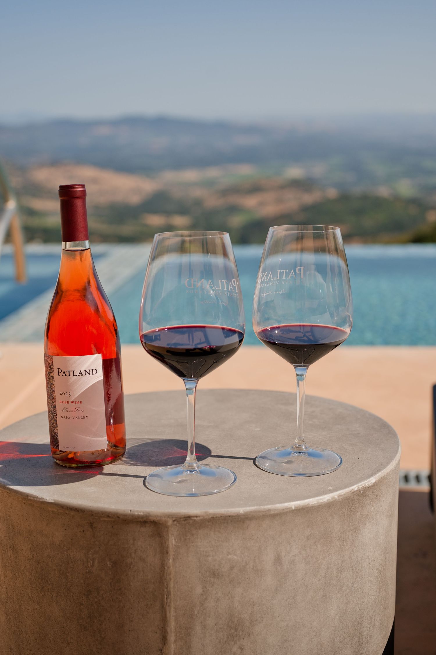 Wine glasses and bottle displayed on concrete ledge with scenic wine country view in background.