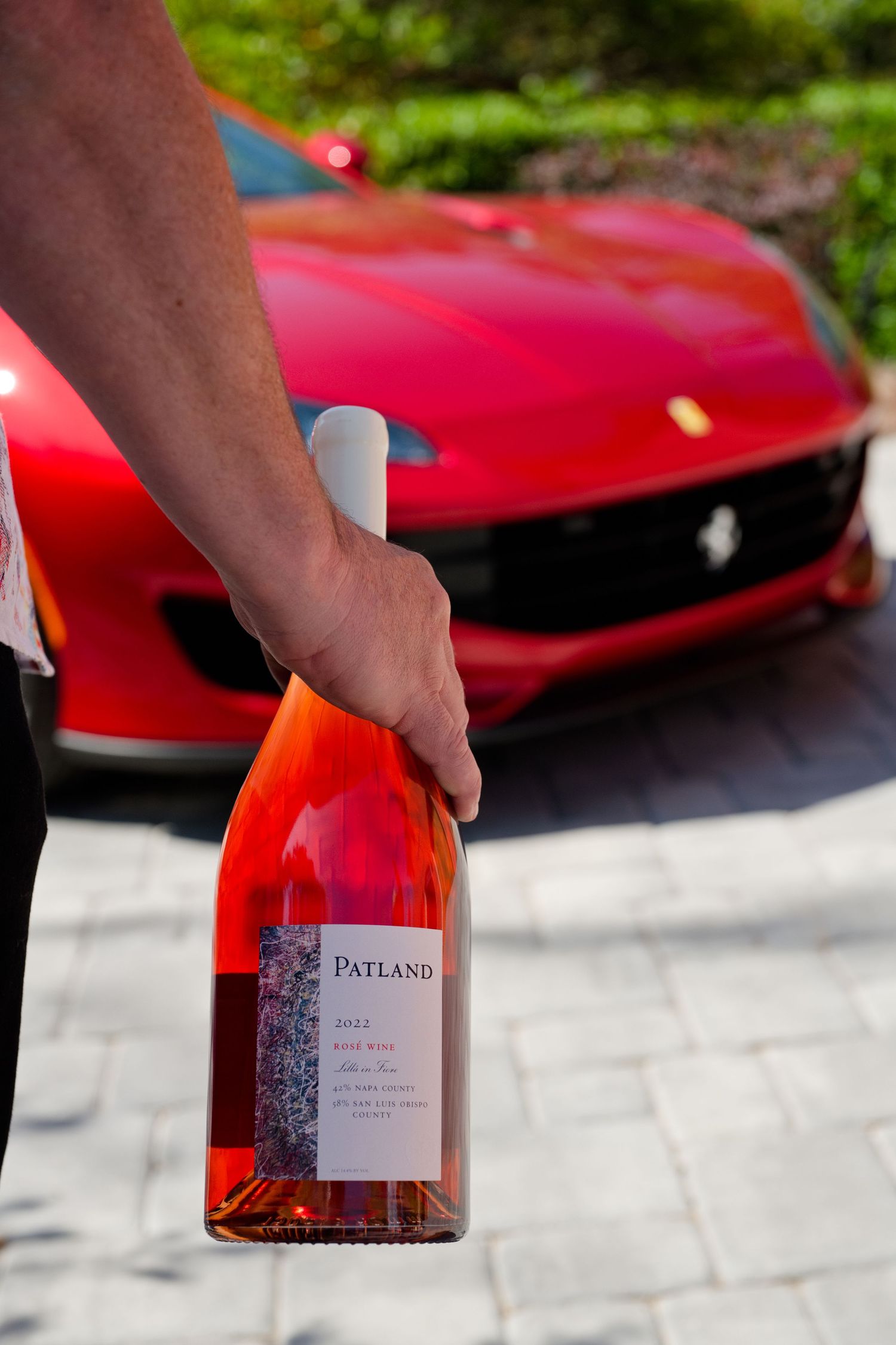 A hand holds a bottle of Patland rosé wine with a red Ferrari in the background, set against the scenic landscape of Napa Valley.