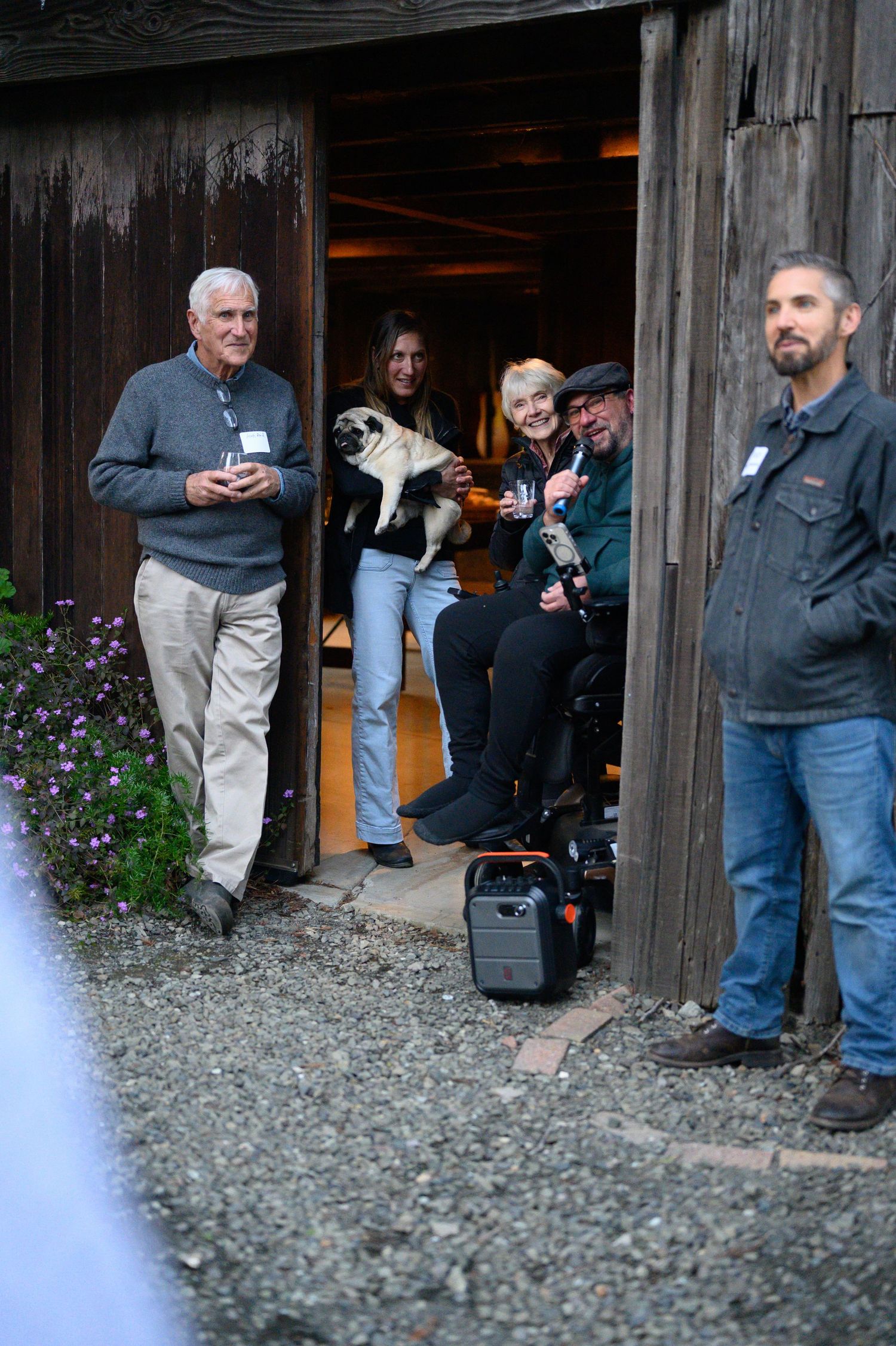 People examine items in rustic outdoor wooden cabin with gravel ground and purple flowers beside doorway.