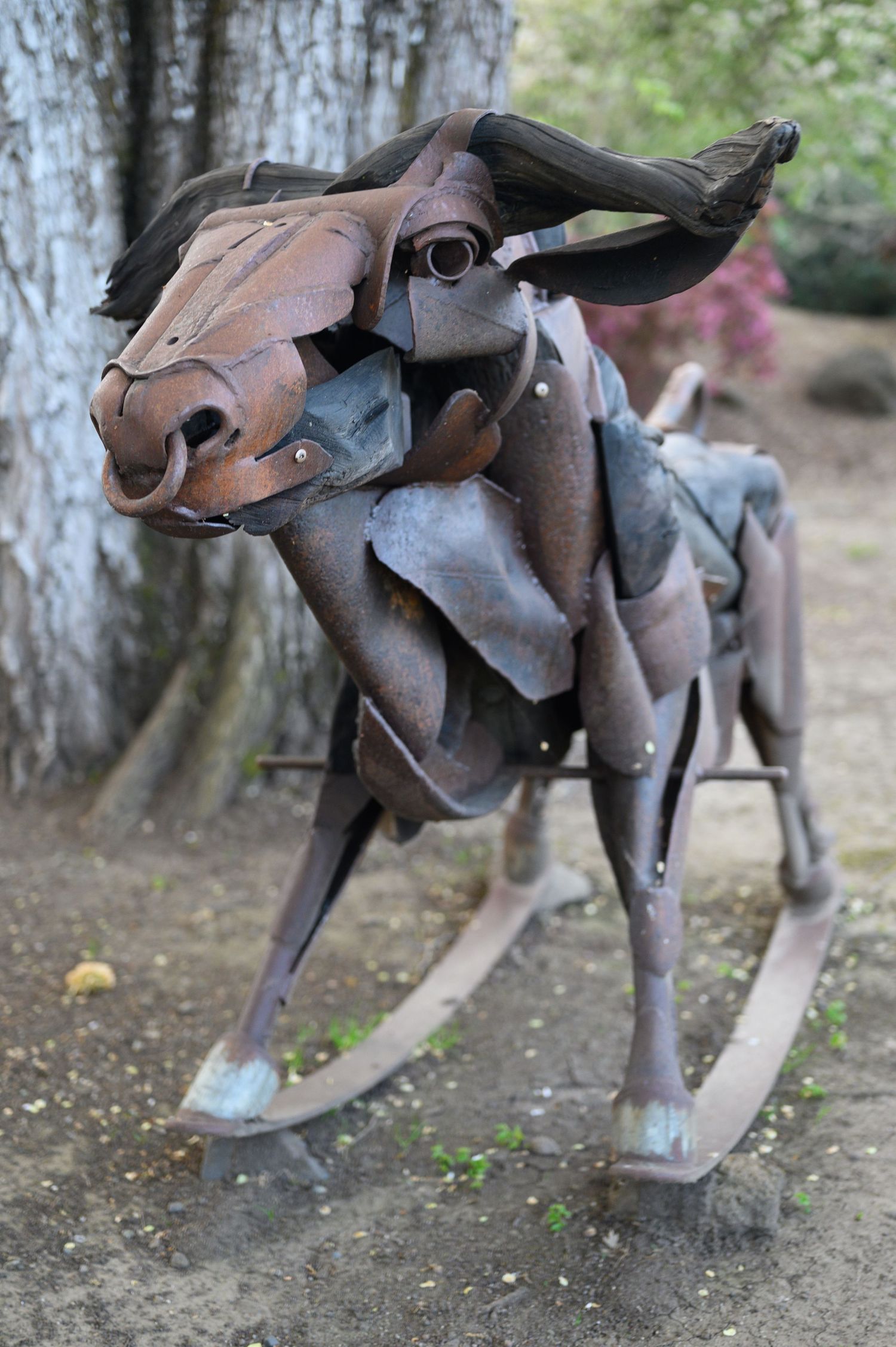 A rustic metal sculpture of a donkey made from repurposed metal parts stands on dirt ground near tree trunks.