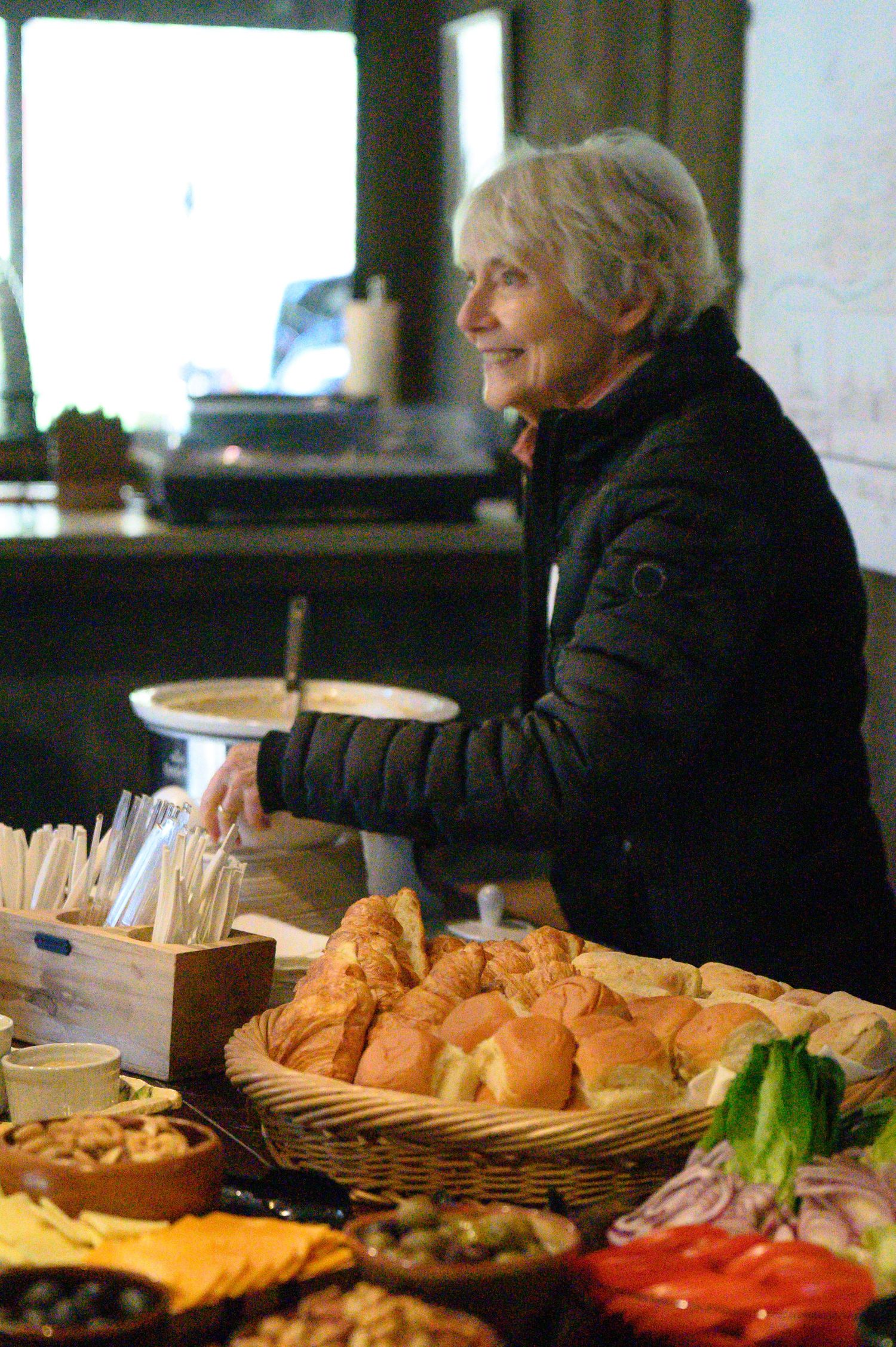 A buffet table with fresh bread rolls, sandwich fixings and condiments being prepared at a food service counter.
