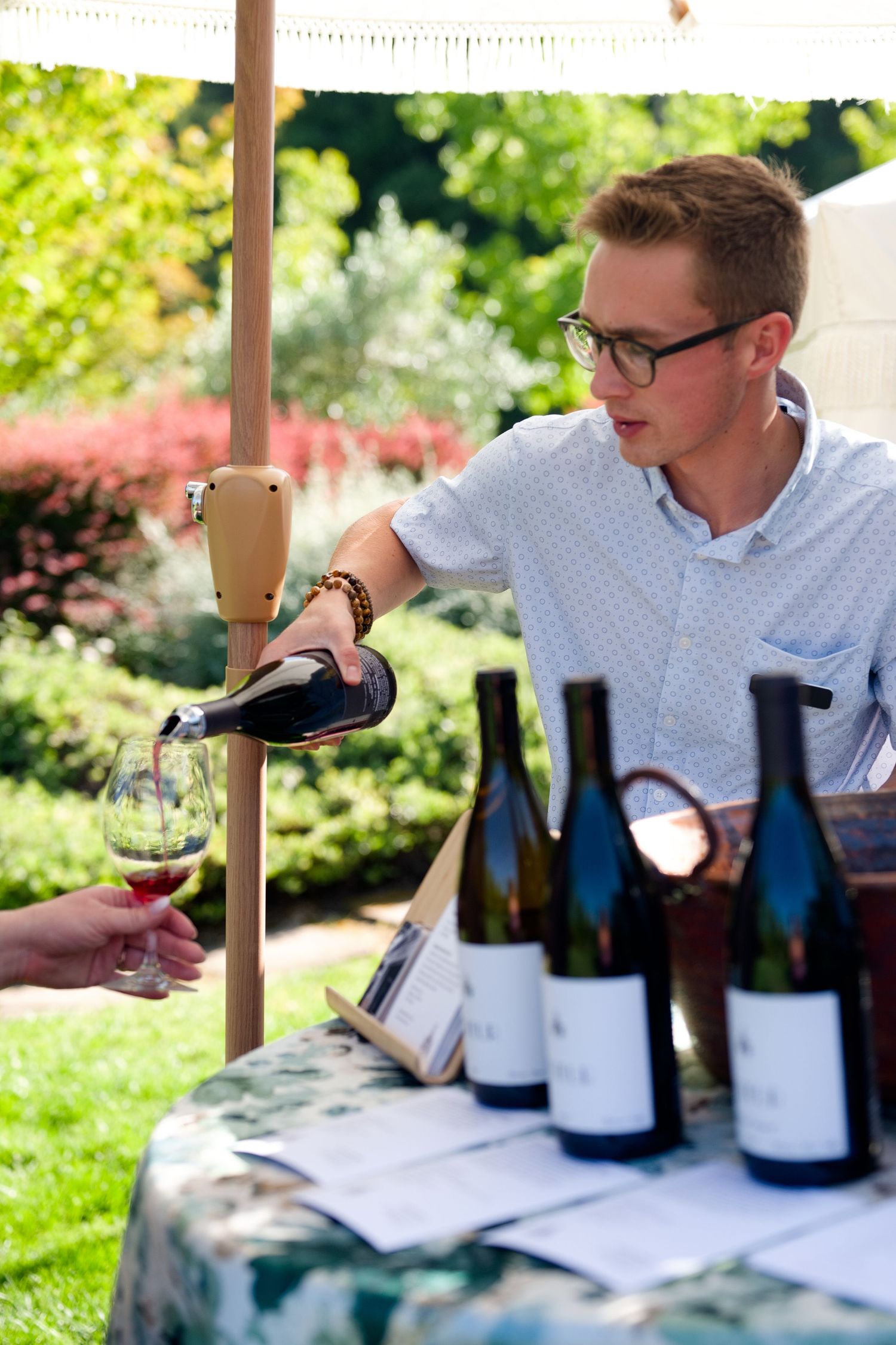 Wine pouring from bottle into glass at outdoor tasting event with wine bottles displayed on table.