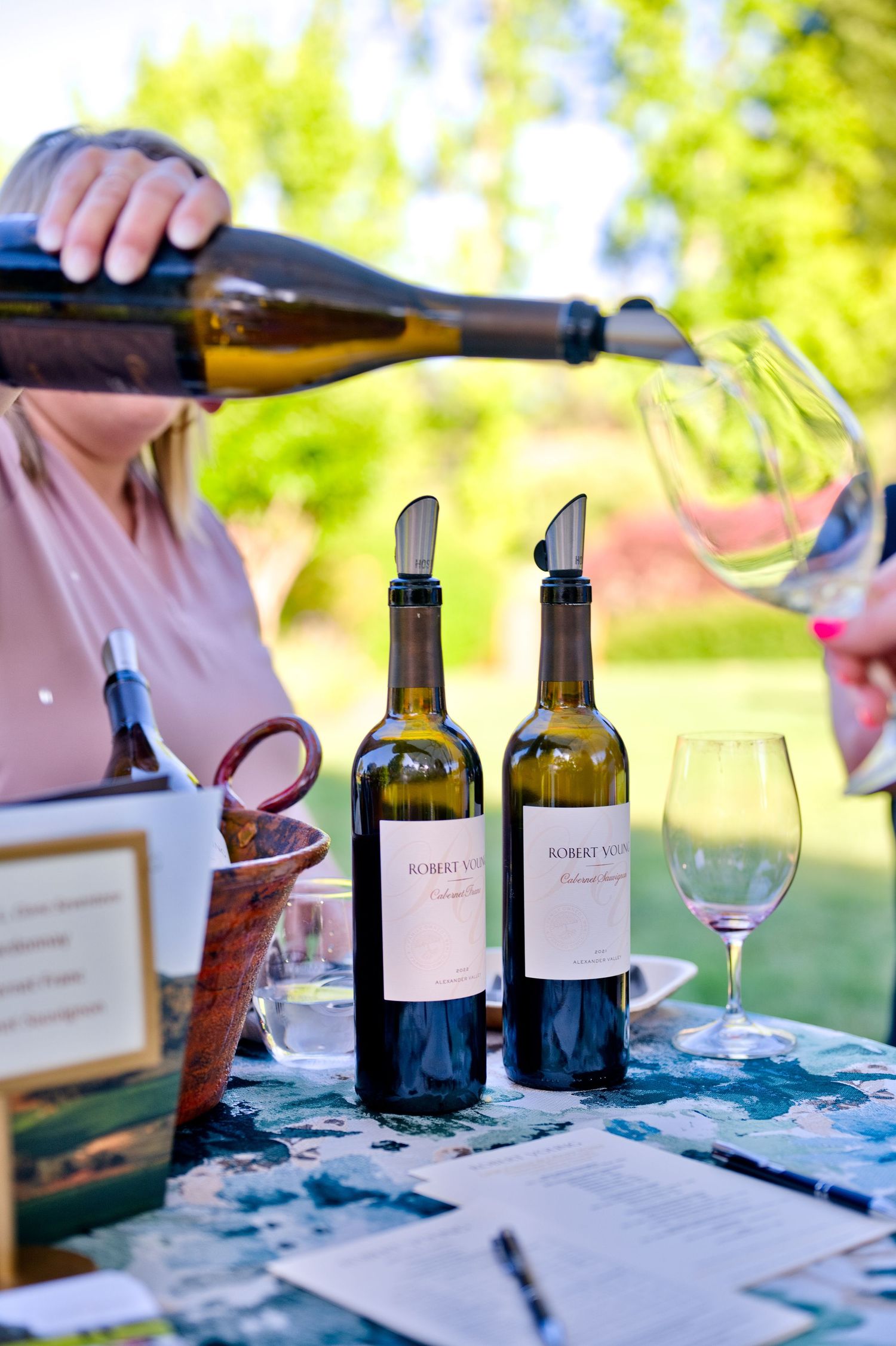 Wine bottles and glasses arranged on outdoor table during summer tasting with sunlit garden in background.