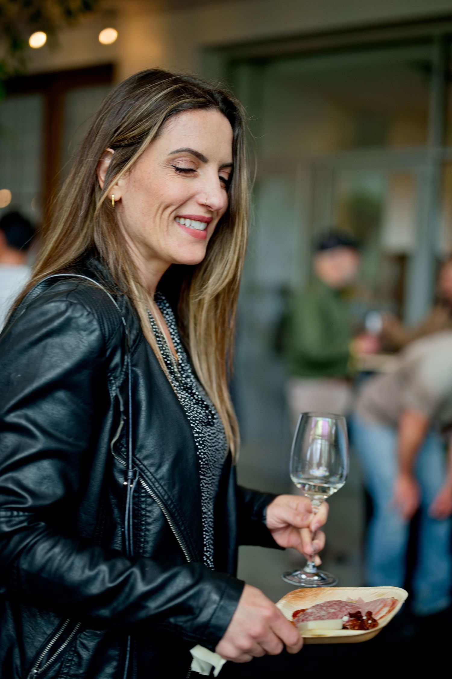 Person wearing a leather jacket smiles while holding a wine glass and plate at a social gathering.