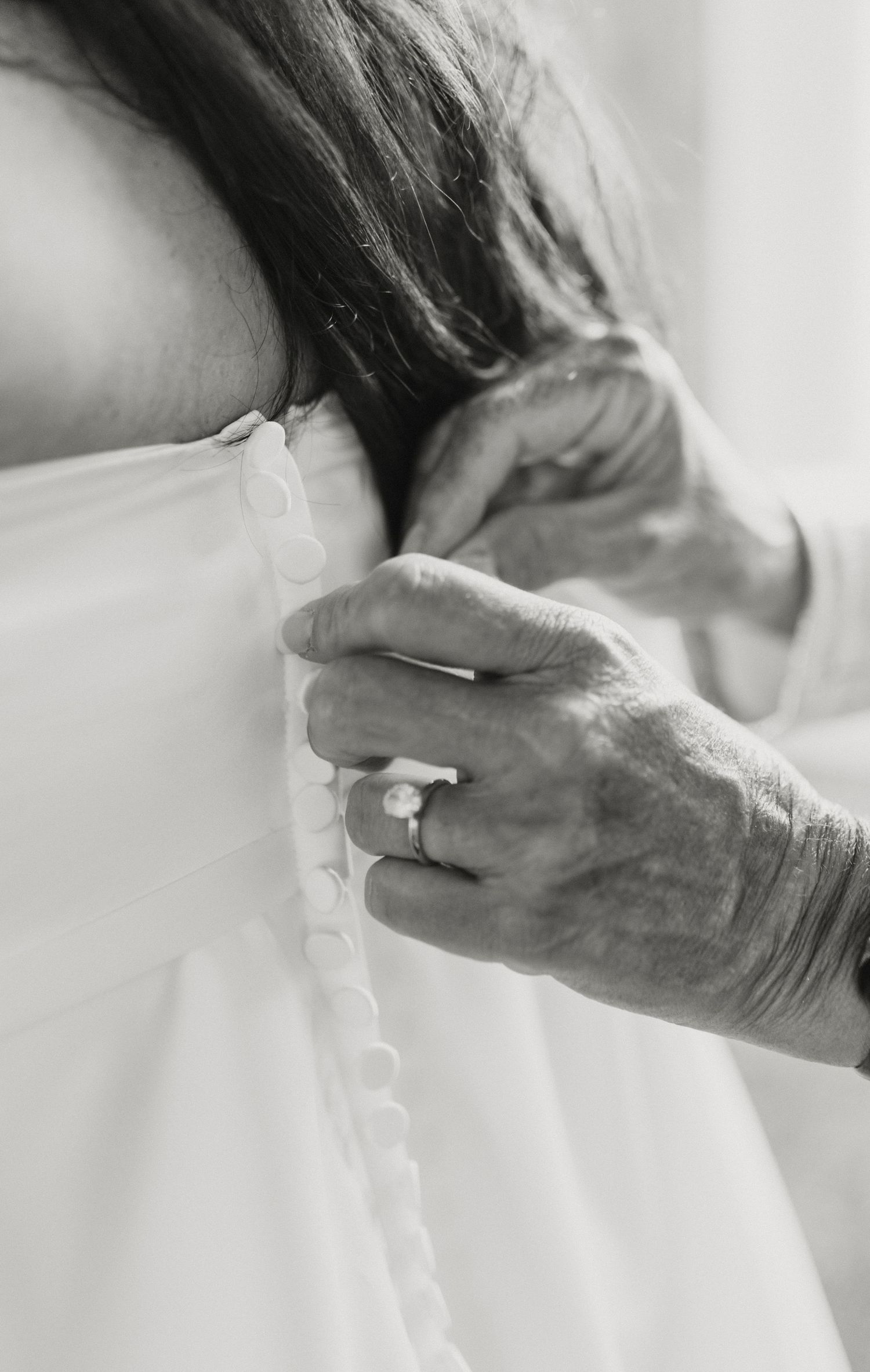 A hand with a wedding ring fastens delicate white buttons on a wedding dress in an intimate black and white moment.