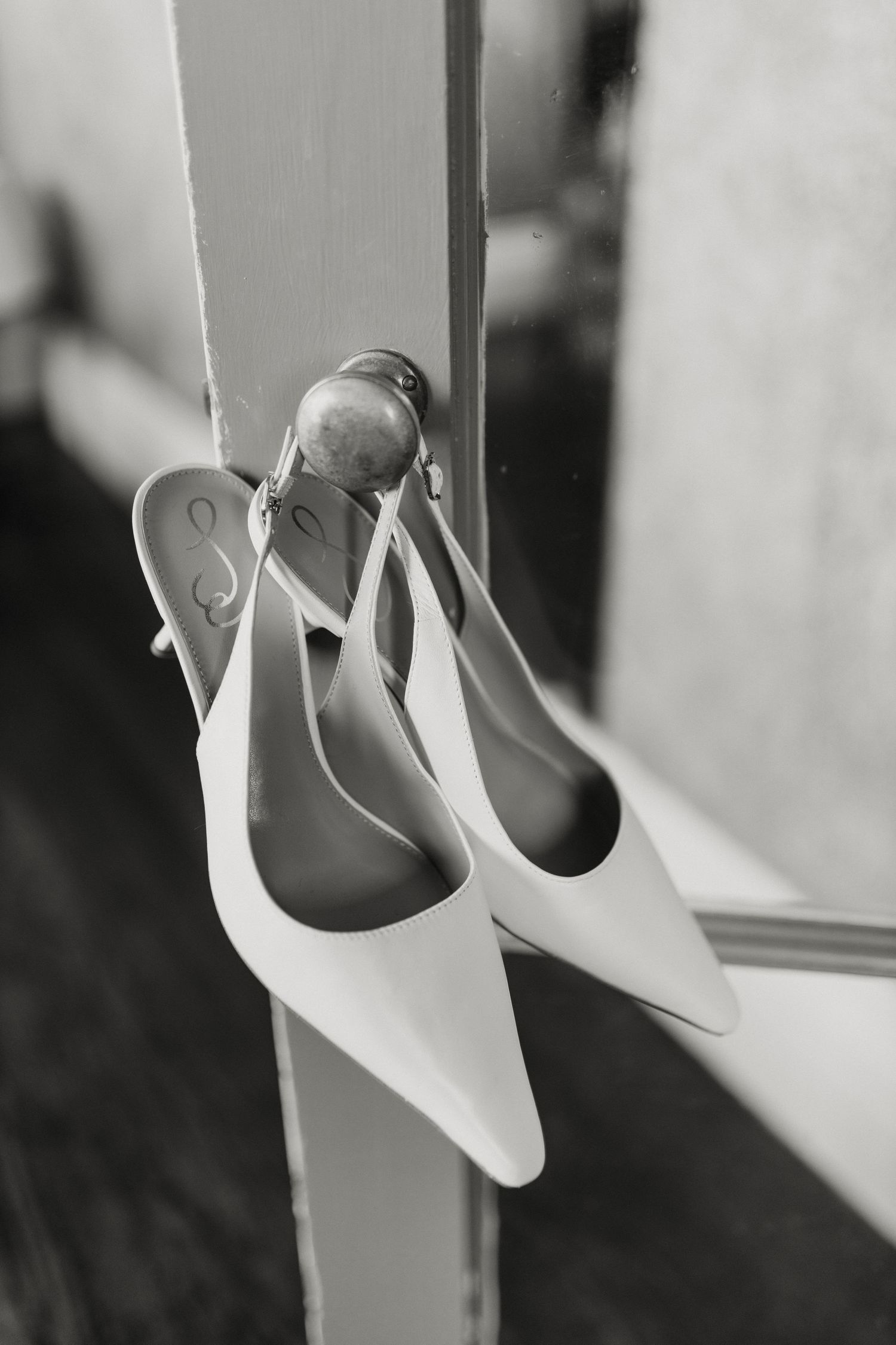 Elegant white bridal slingback shoes hanging on a door frame in black and white photography.
