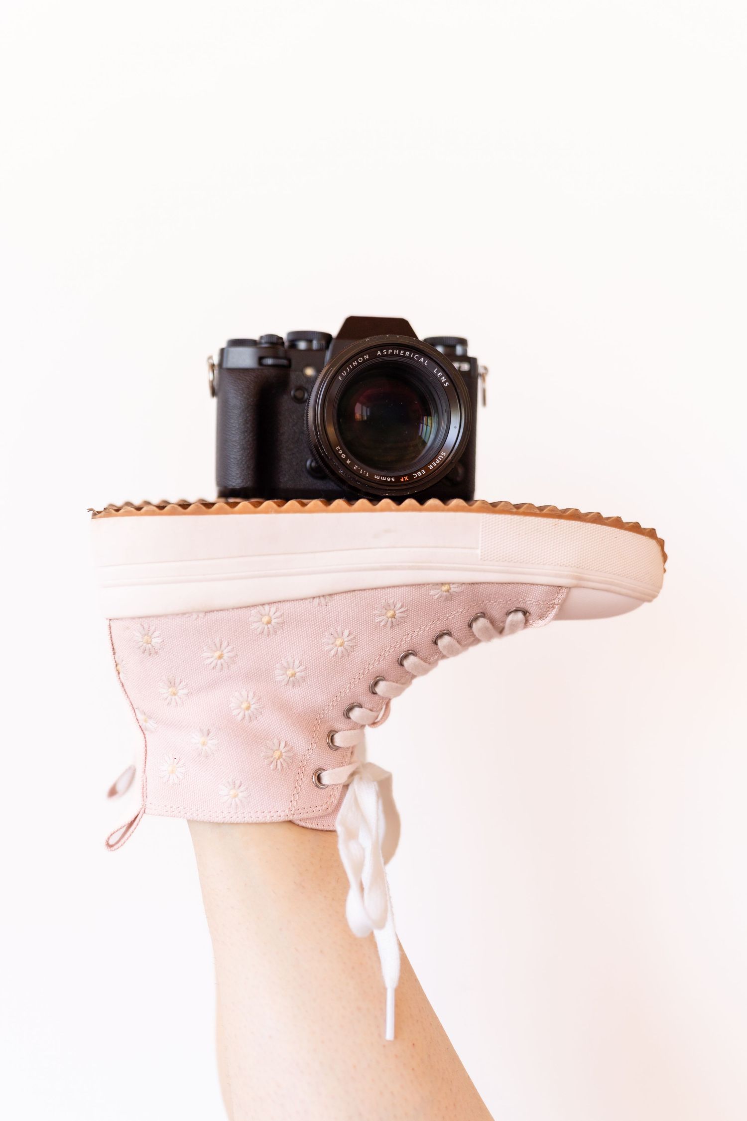 Black digital camera perched on top of a light pink high-top sneaker against white background.