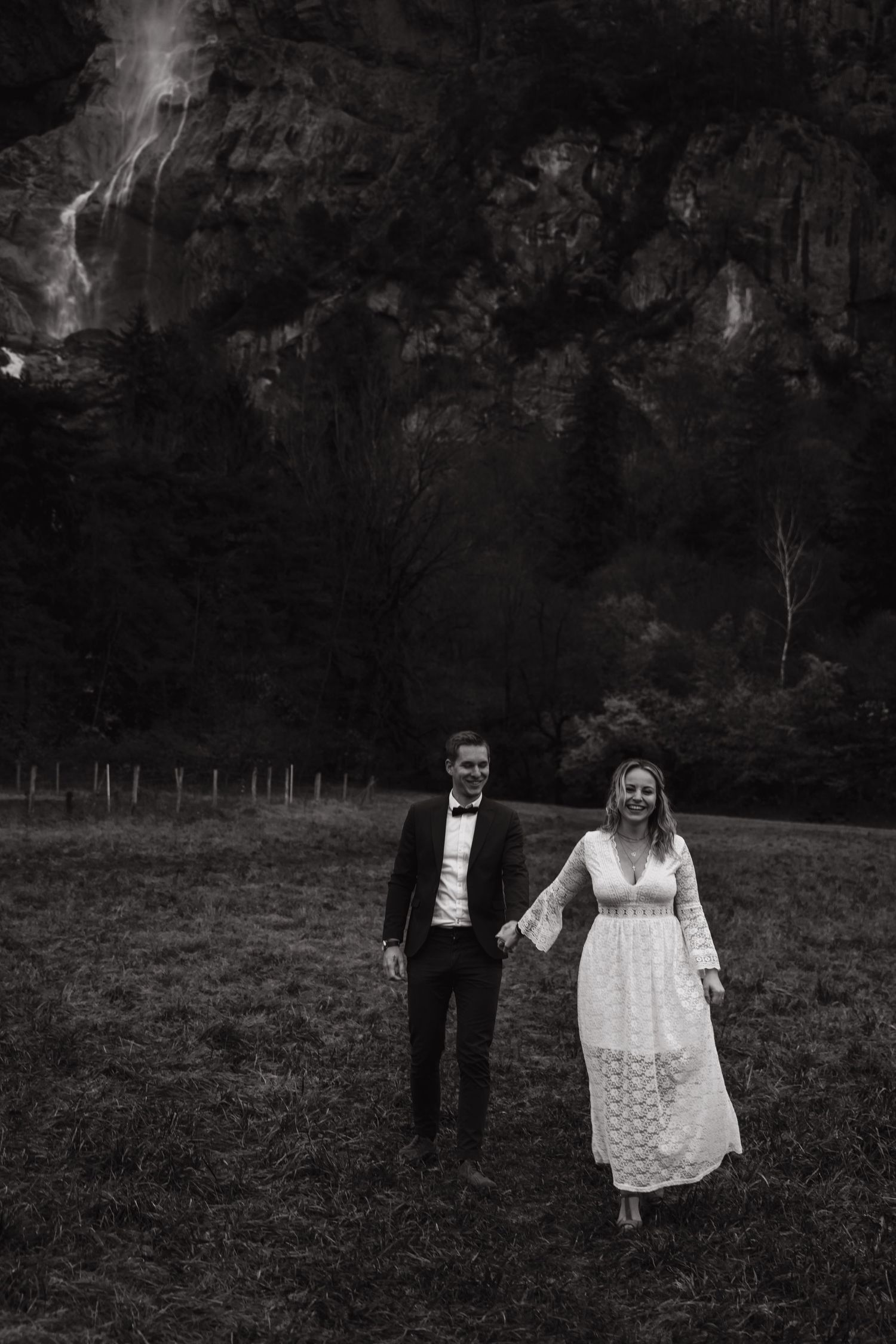 A black and white photo of newlyweds walking hand in hand in a field with dramatic cliffs in the background.