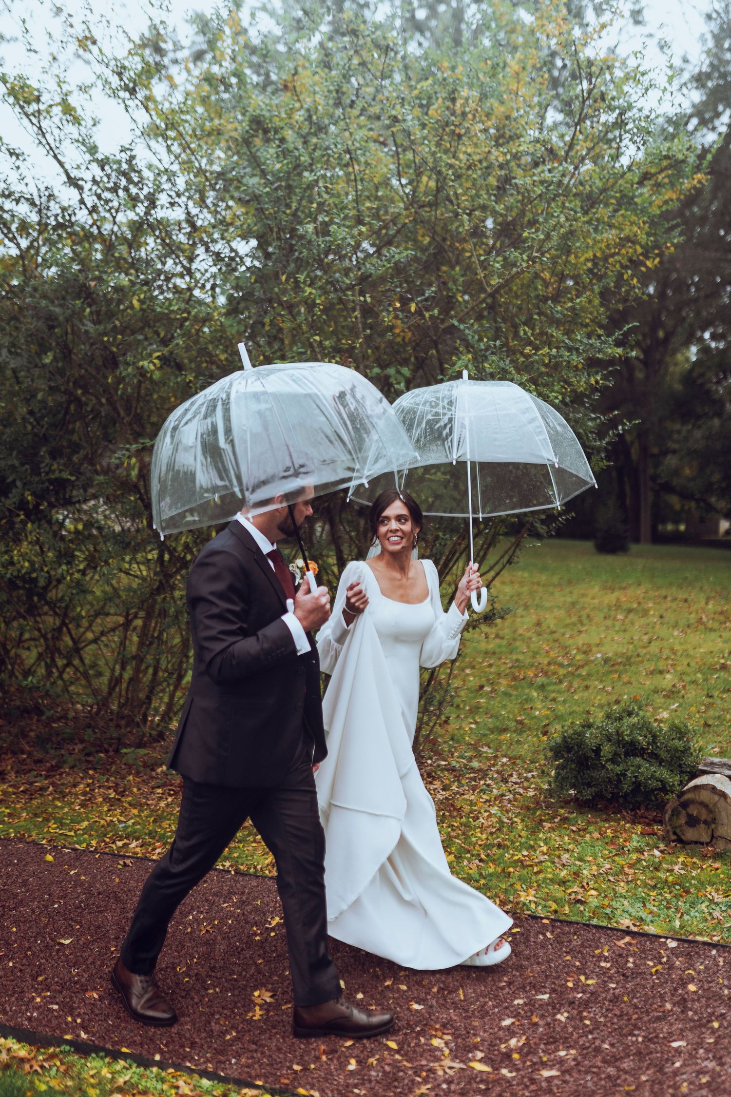 A bride and groom walk hand in hand in the rain, smiling under clear umbrellas, capturing a romantic and joyful wedding moment.