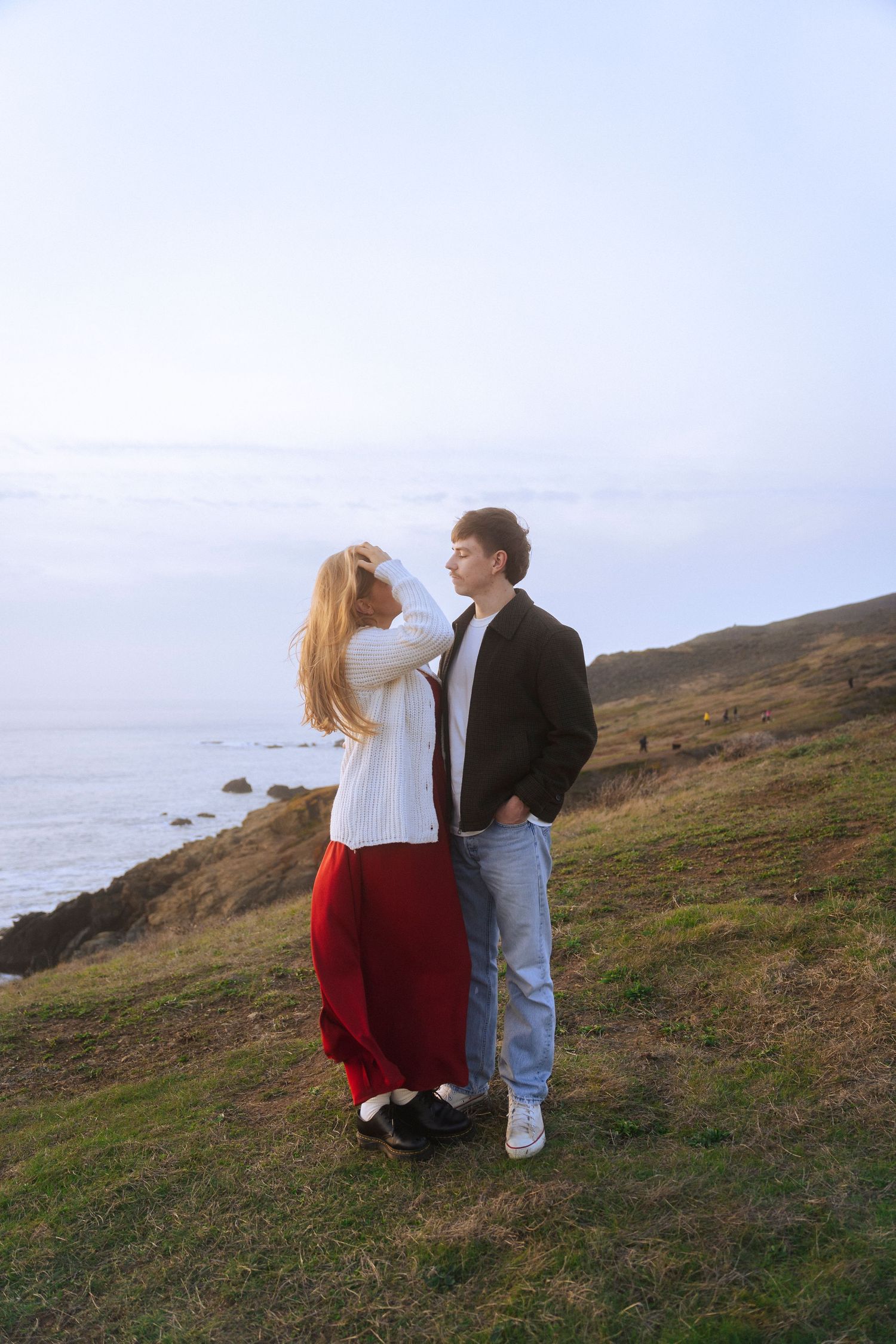 A couple stands on a sandy beach at sunset, holding hands, with green hills behind them. A warm, romantic moment captured in golden light.