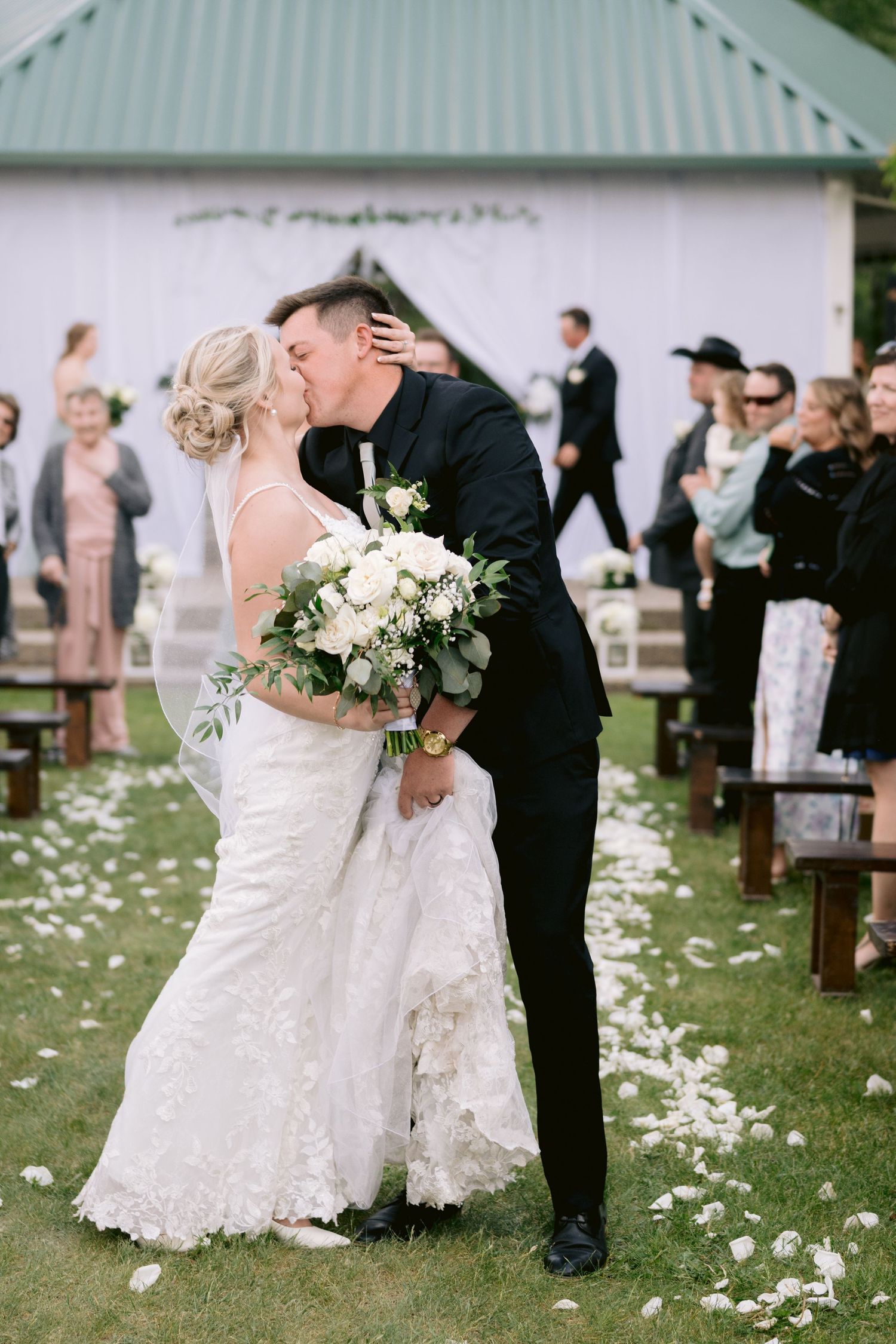 White petals are scattered on green grass around elegantly dressed wedding guests standing near wooden benches.