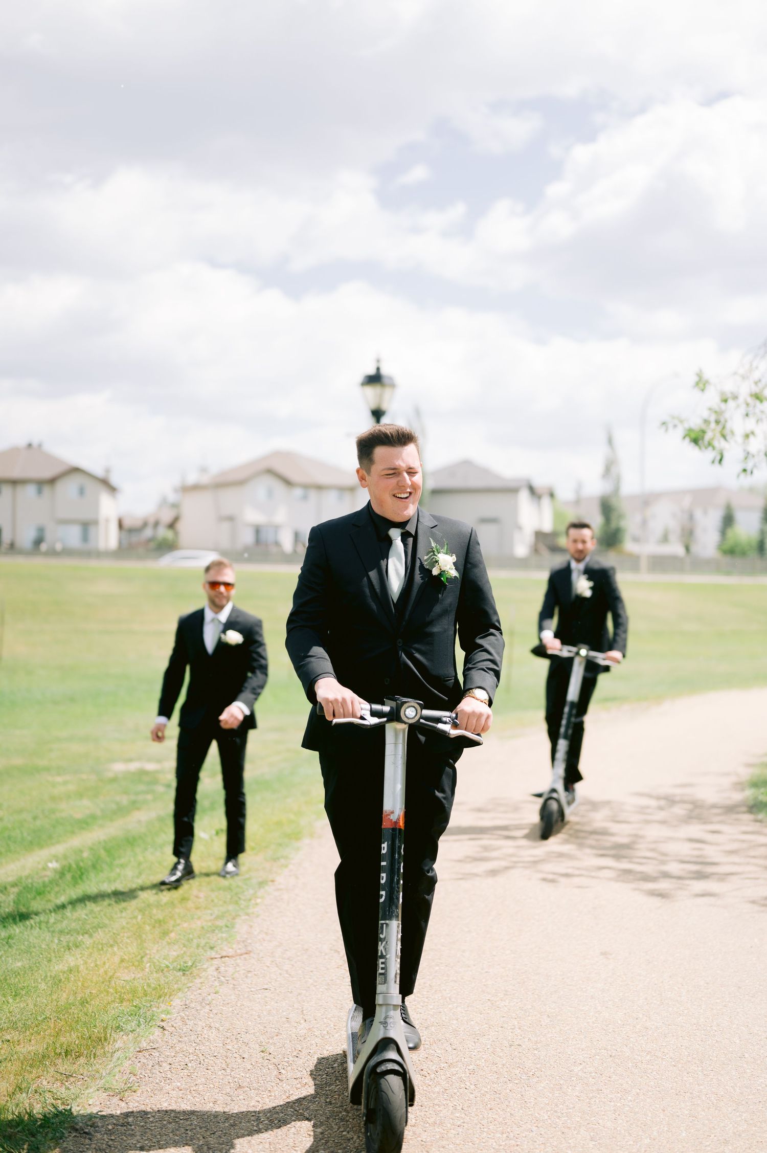 Groomsmen in black suits walk confidently down a path during wedding day preparations.