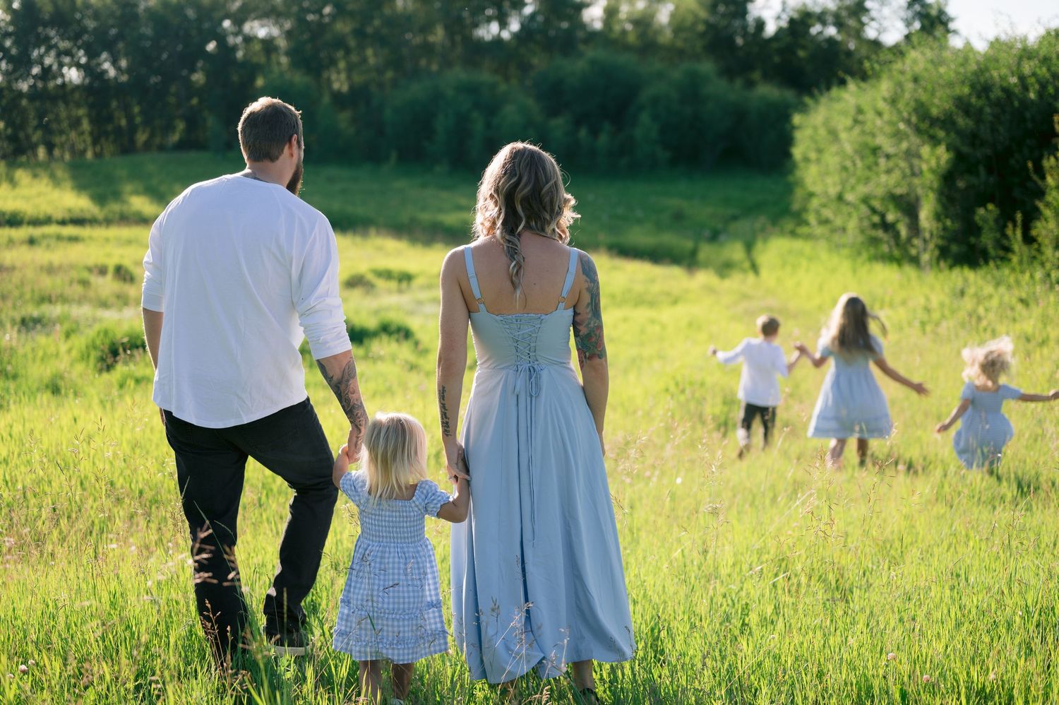 A family walks together in a green summer meadow at sunset while children play in the distance.