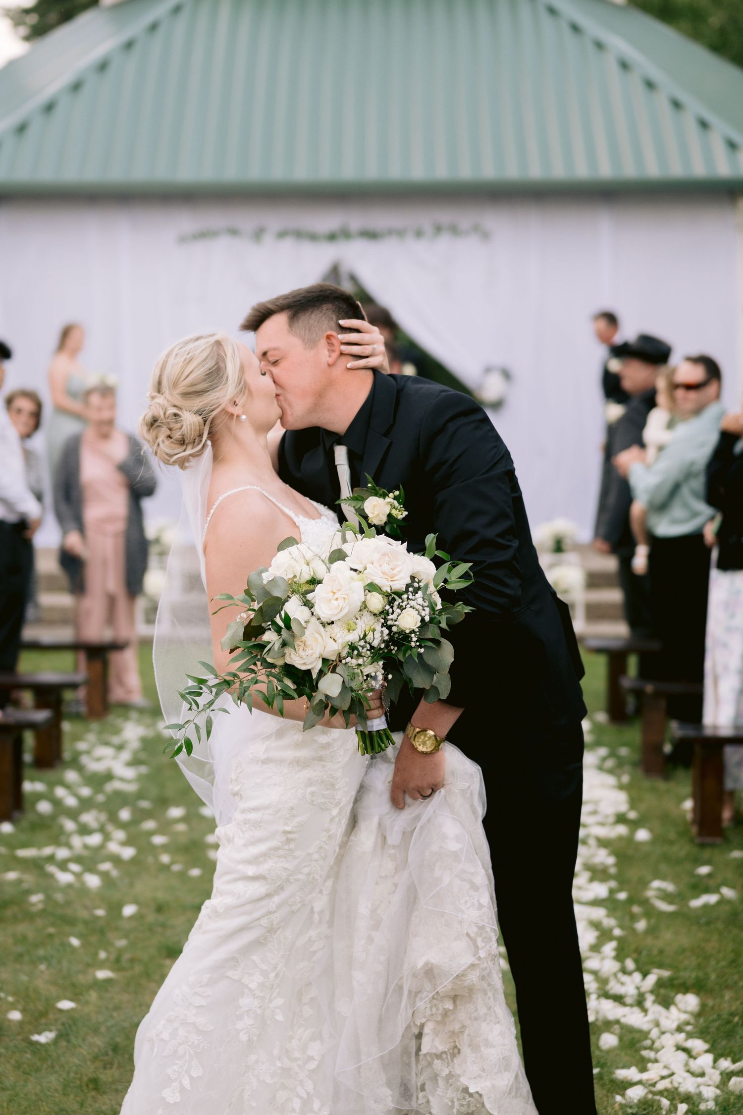 A bride and groom share their first kiss during an outdoor wedding ceremony with white flower petals.