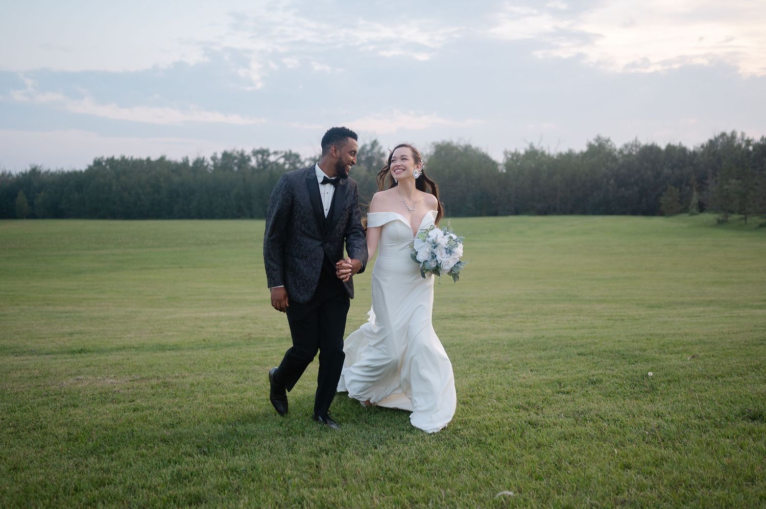 Couple runs playfully through a grassy meadow at sunset during their wedding day.