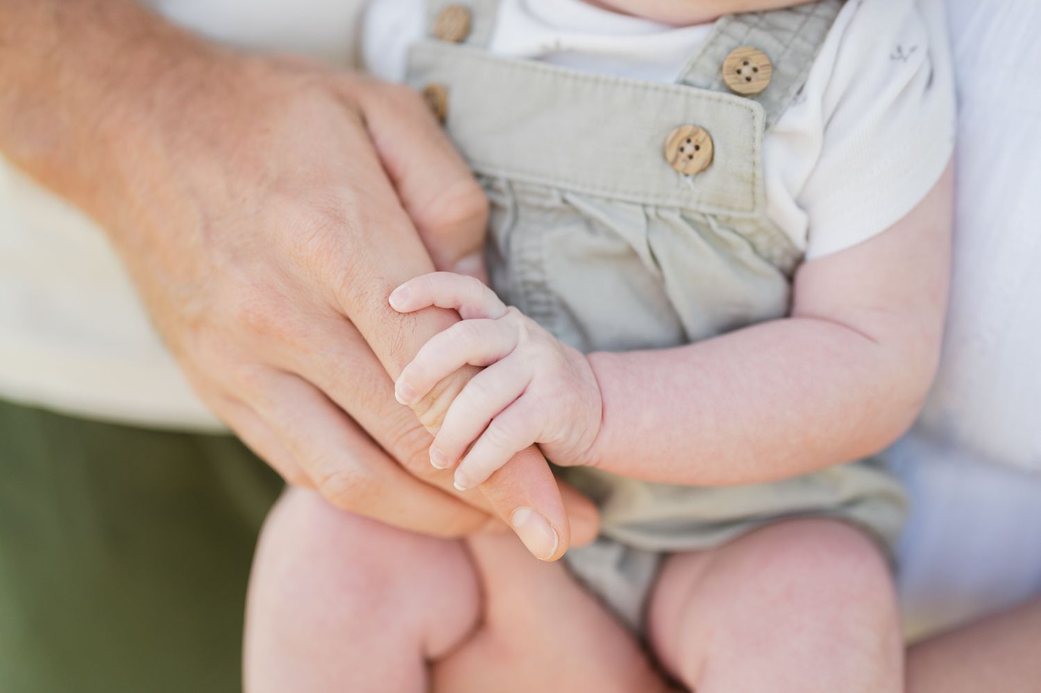 A close-up of tiny baby fingers gripping an adult's hand, with the baby wearing light gray overalls.