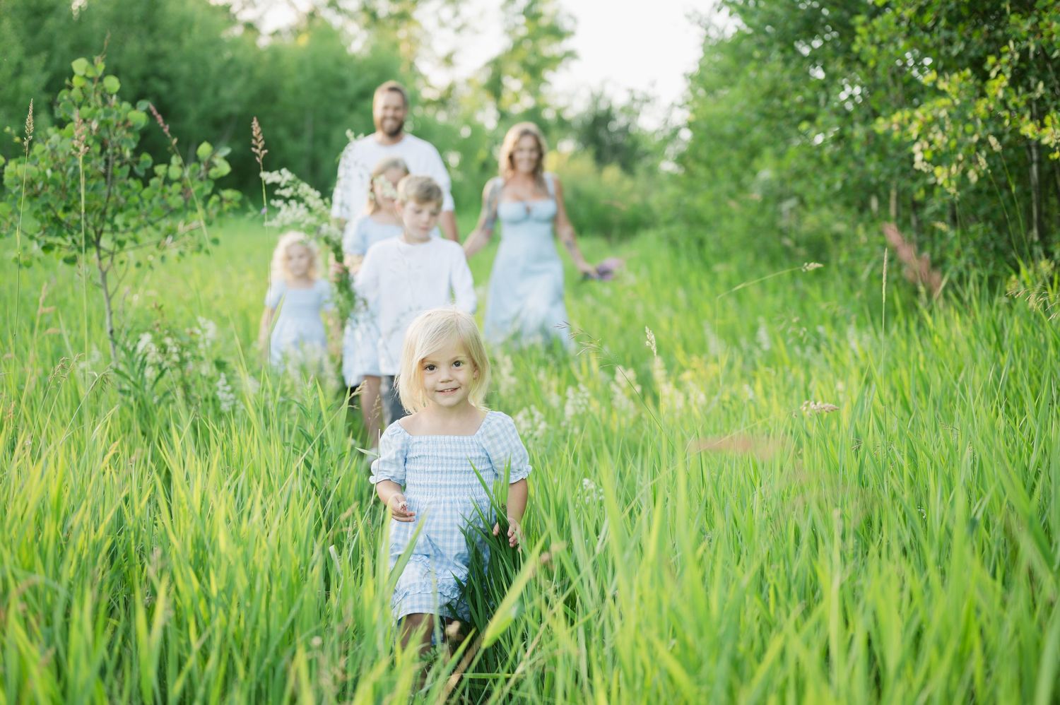 A family in light blue and white outfits runs joyfully through a lush green meadow on a sunny summer day.