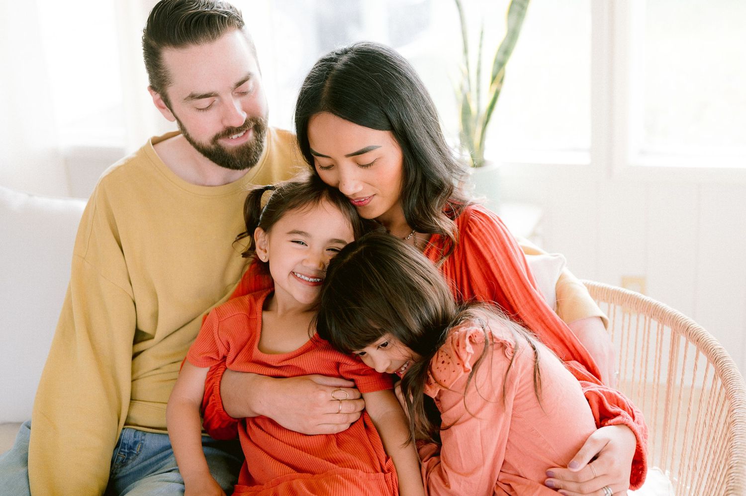 A happy family shares a warm embrace while sitting together on a pink chair in a bright, sunlit room.