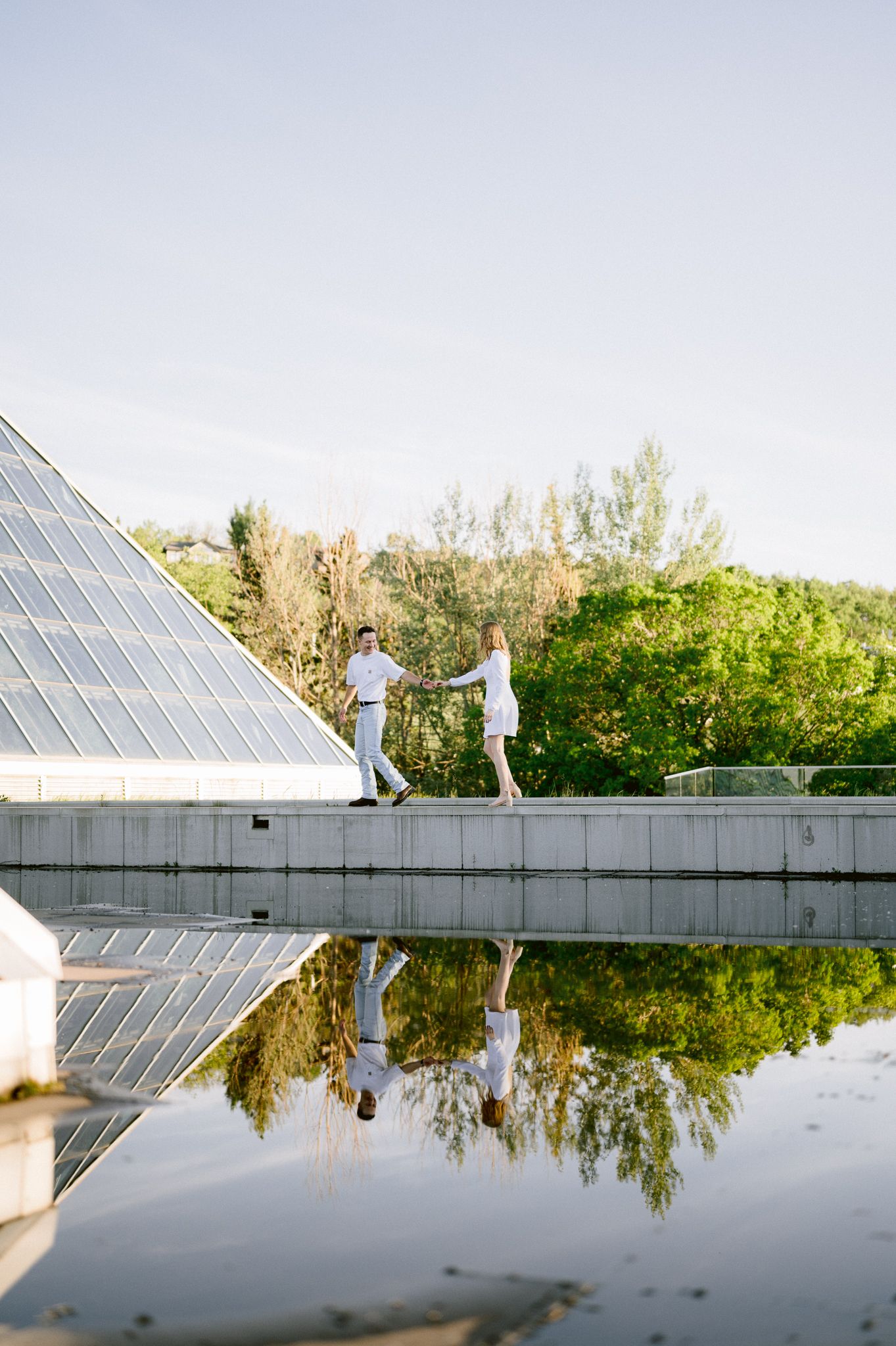 A couple walks along a modern glass structure with their reflection in the water below.