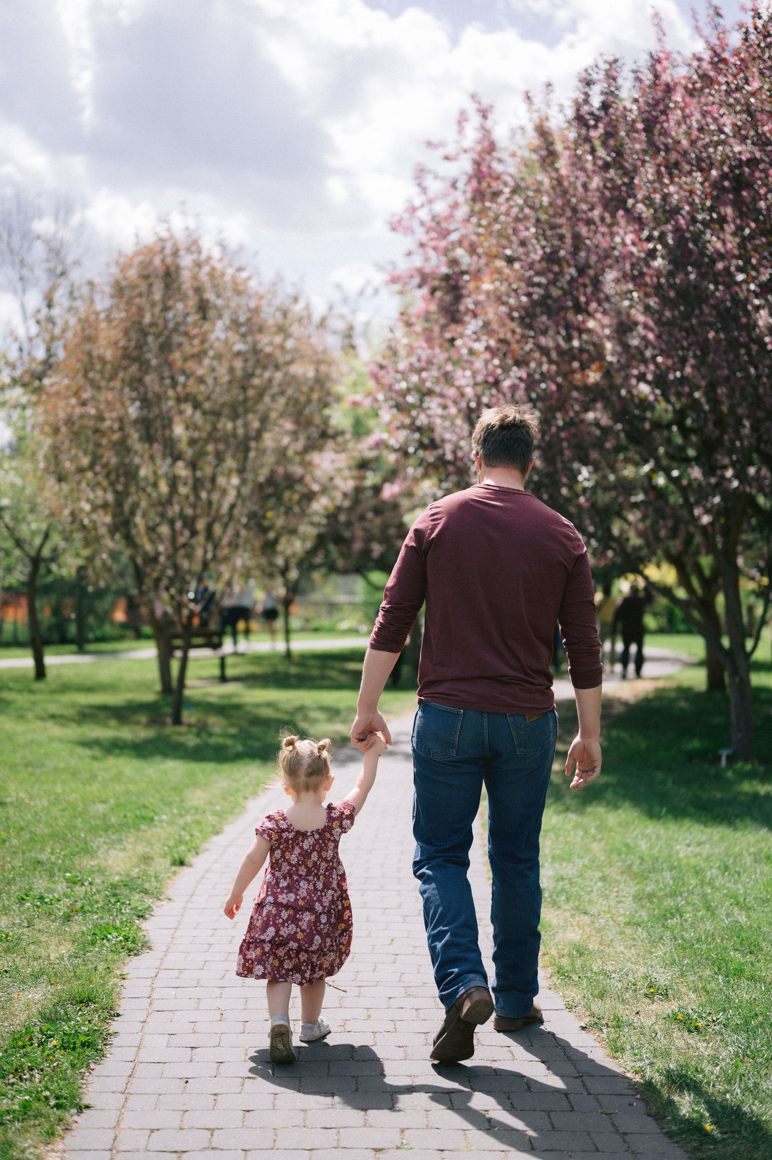 Father and daughter walking hand in hand on brick path through spring blossoms.