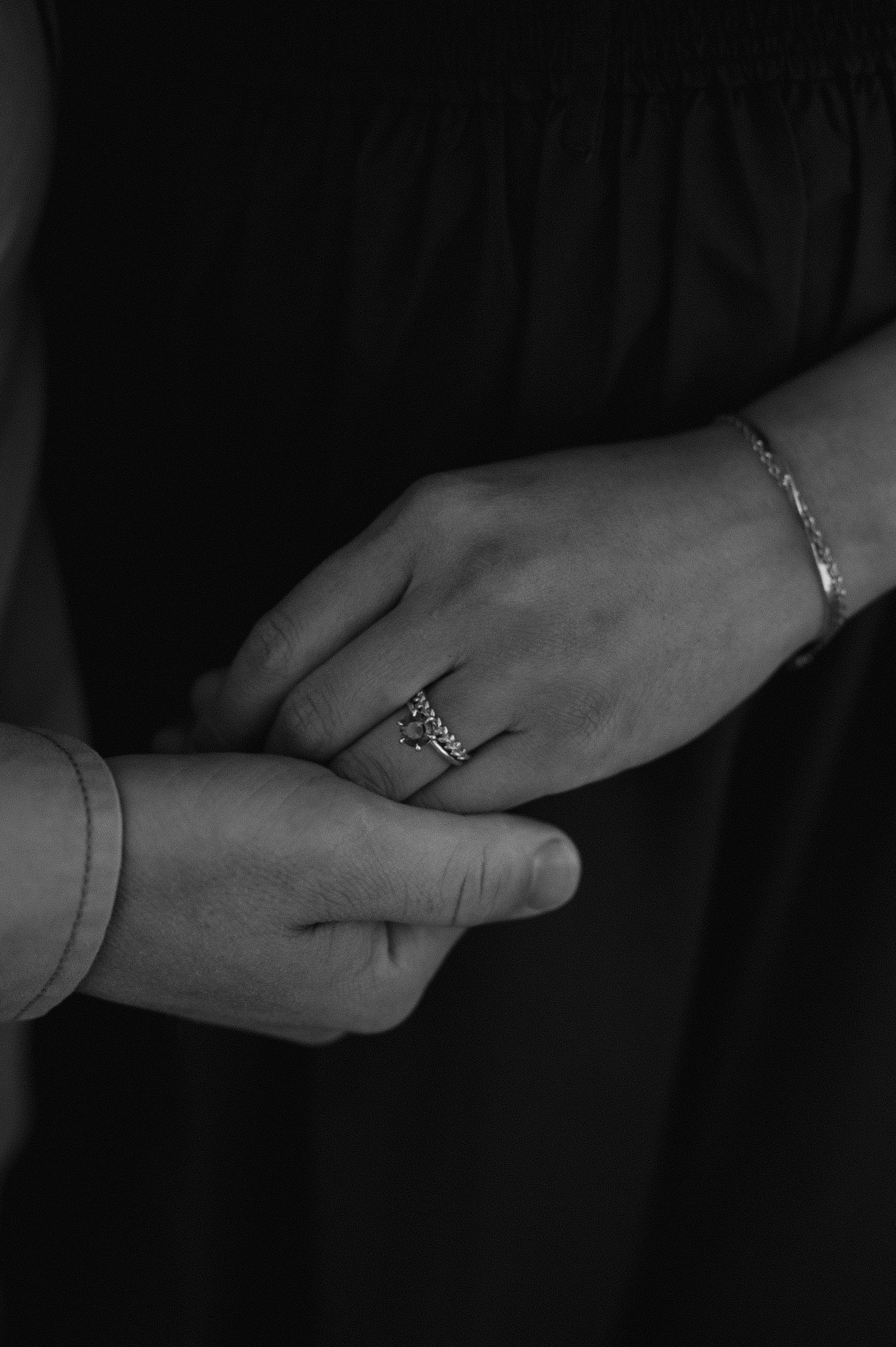 Black and white close-up of hands with wedding rings and a delicate bracelet against a dark background.