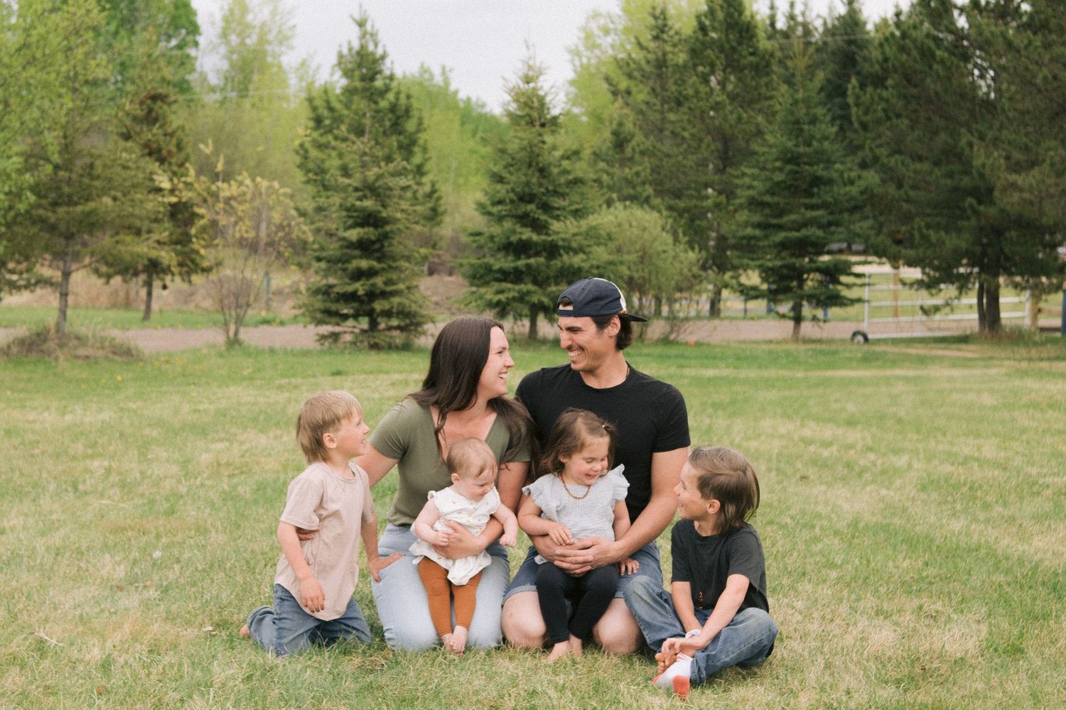 Family sitting together on grass in a park surrounded by evergreen trees.