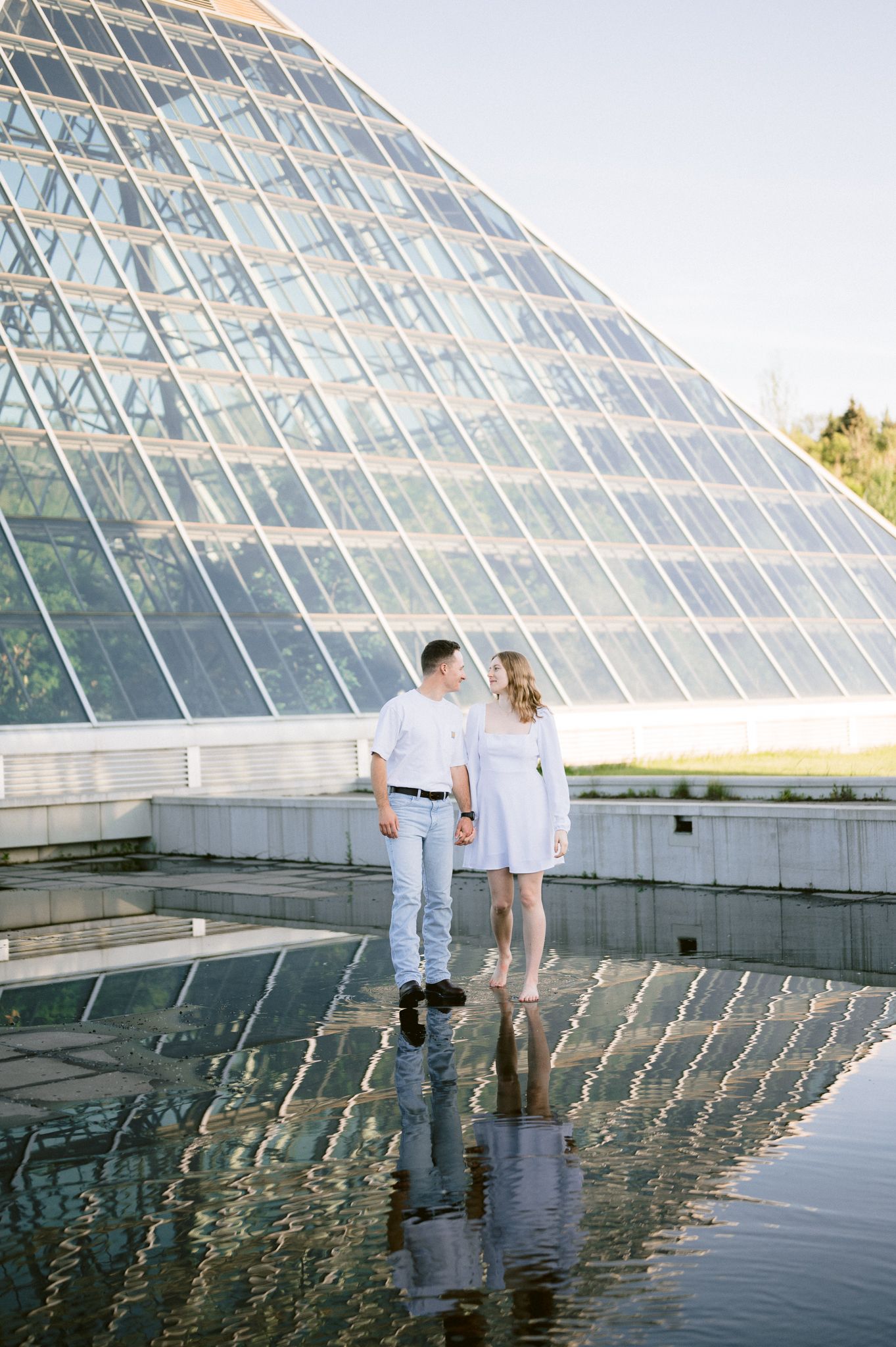 A couple poses by a geometric glass pyramid building with its reflection in the surrounding water.