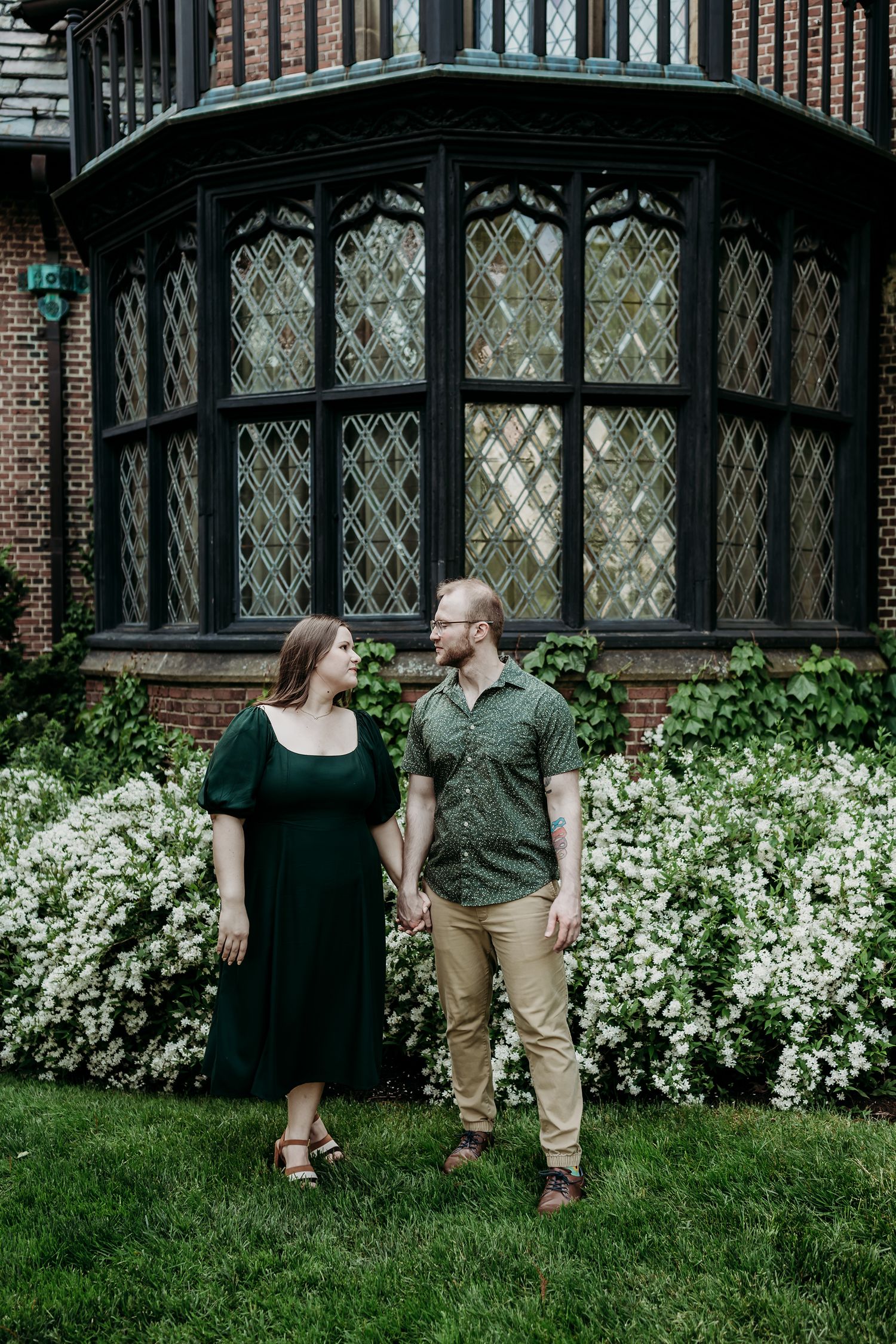 Two people stand smiling together in front of a grand bay window with flowering bushes on a green lawn.