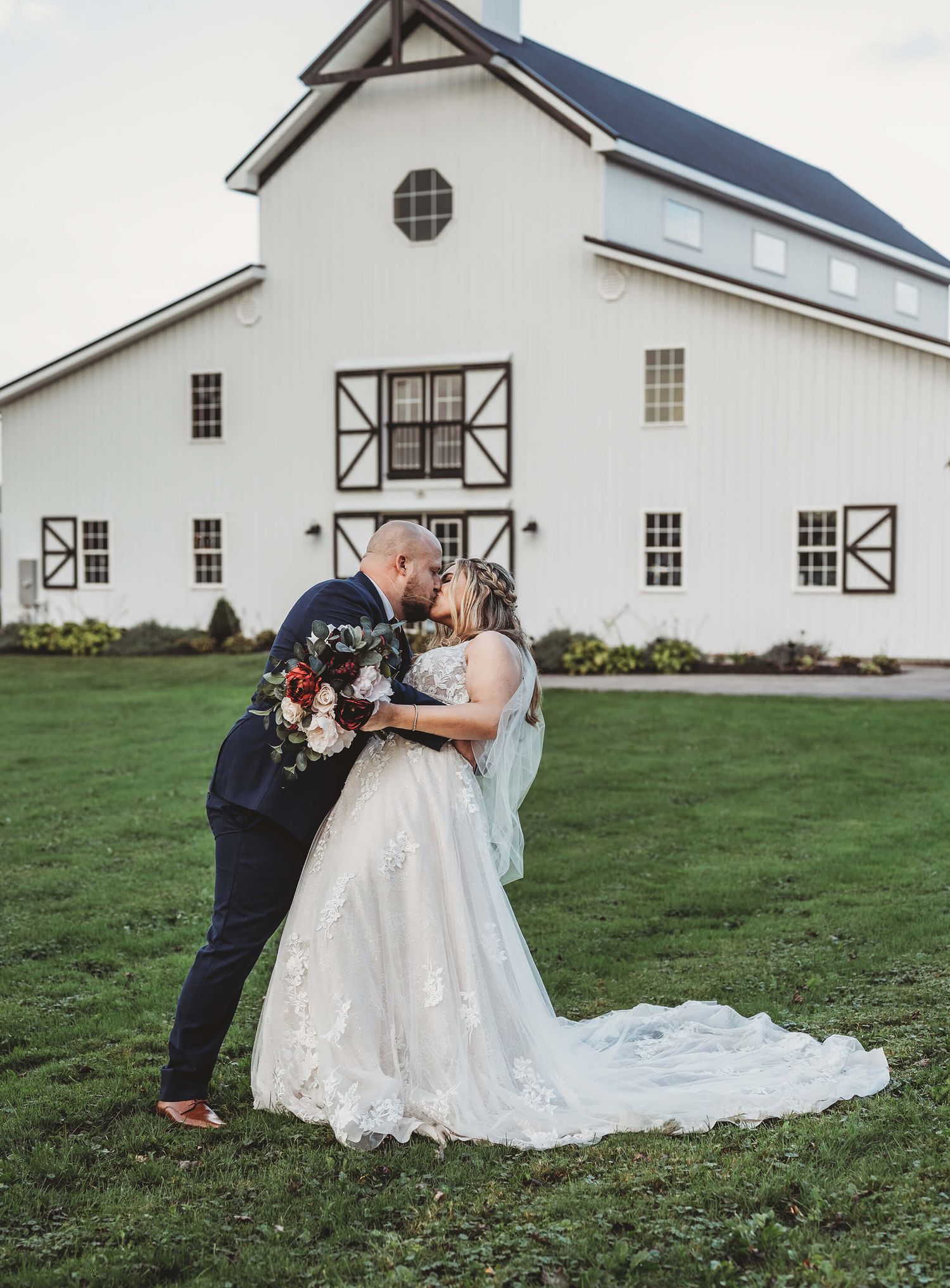 A couple shares a romantic kiss on their wedding day in front of a white barn-style venue with black trim.