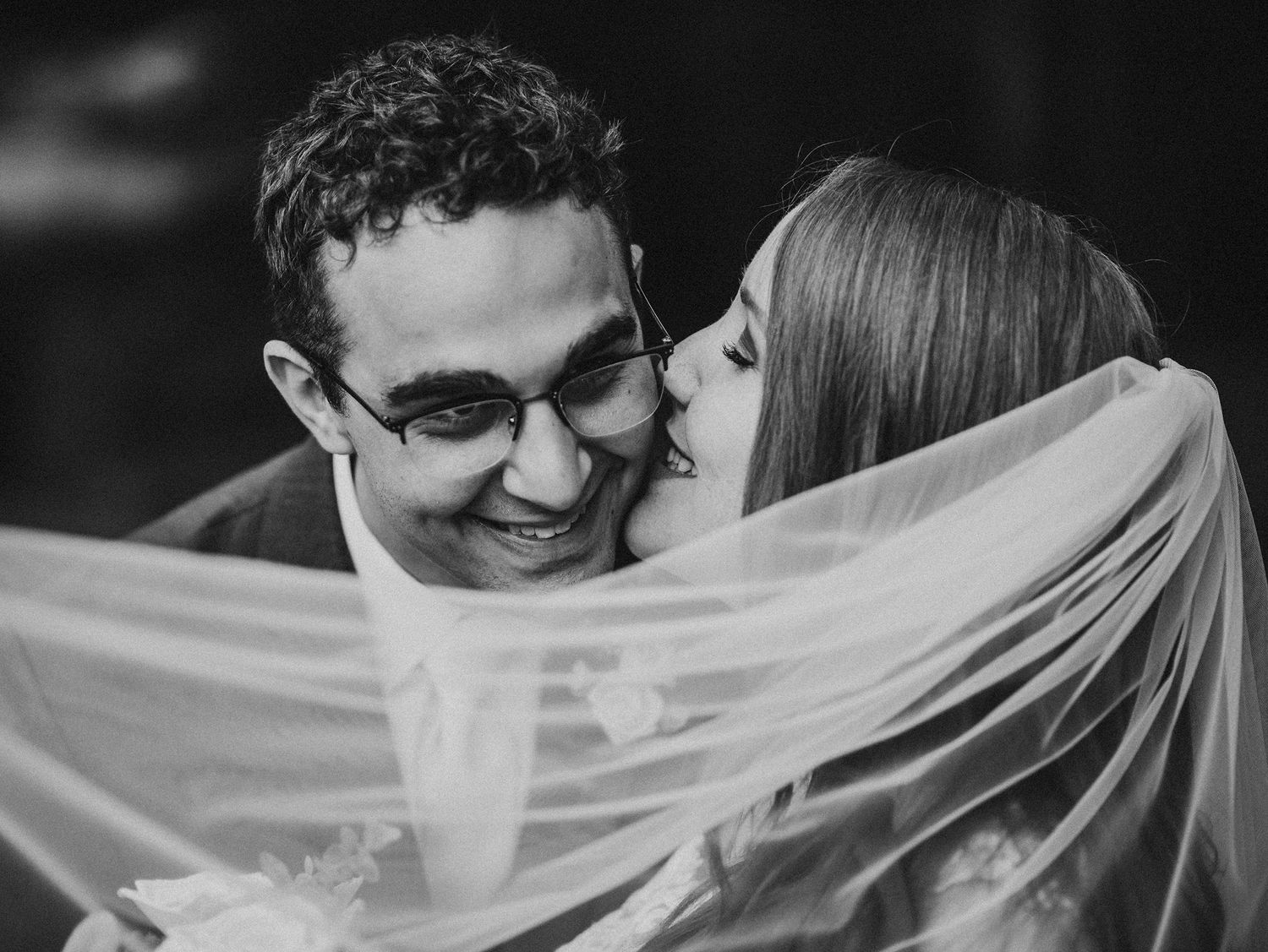 Black and white wedding photo showing couple embracing under a flowing veil in an intimate romantic moment.